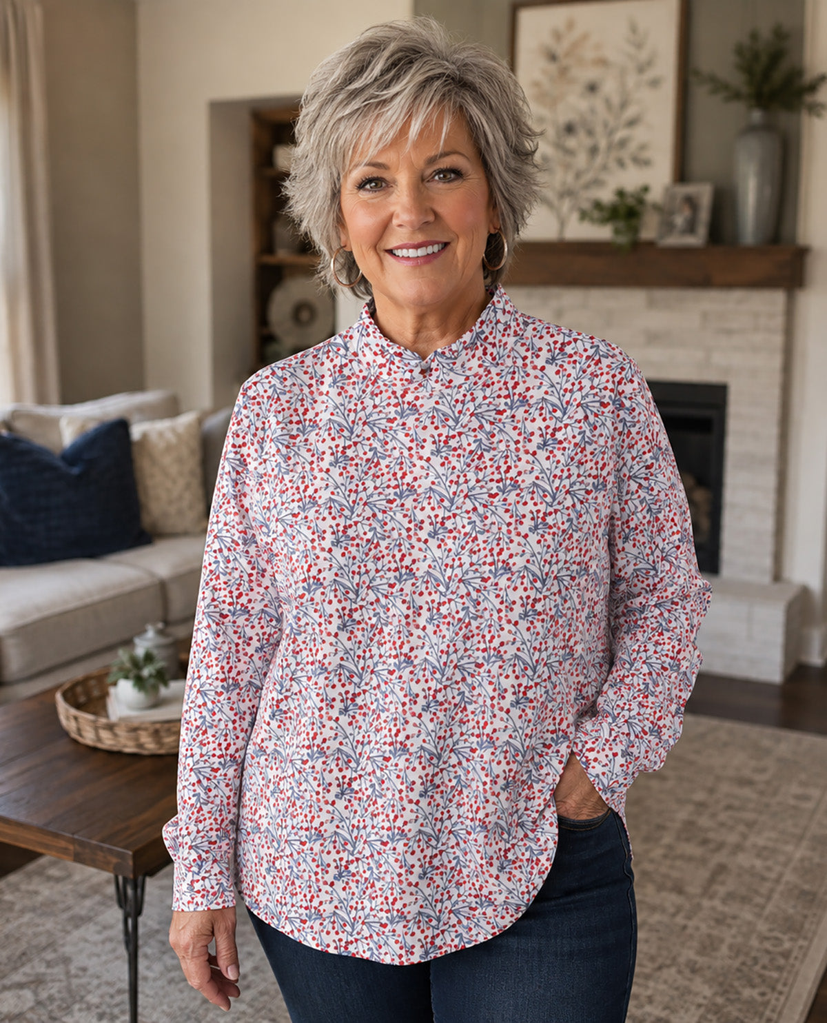 A smiling woman with short gray hair stands in a living room wearing a long-sleeve button-up shirt with a dense red, blue, and white floral print, paired with dark blue jeans.