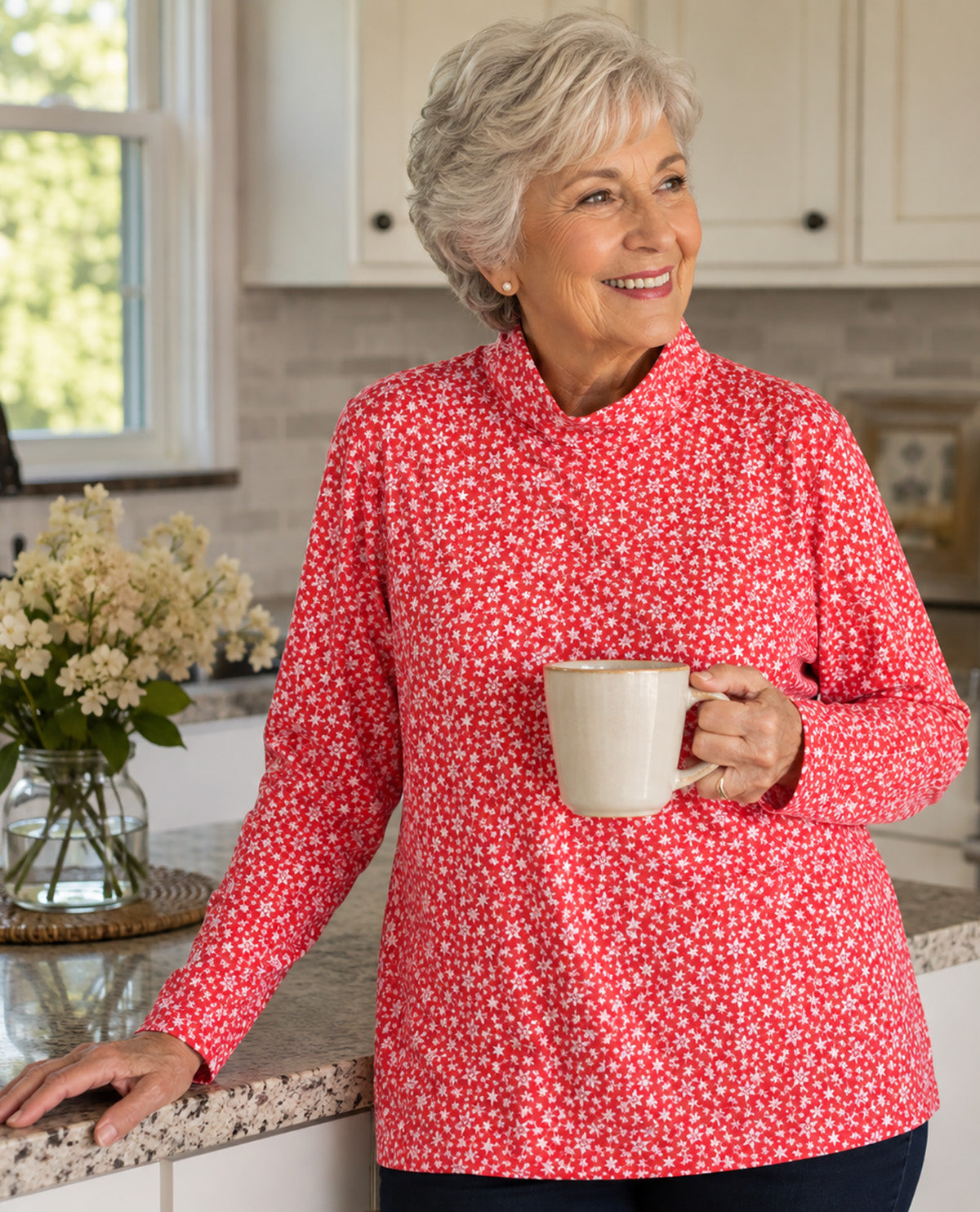 An older woman with short gray hair stands in a bright kitchen, smiling and holding a cream mug. She is wearing a red long-sleeve top with a dense white floral pattern.