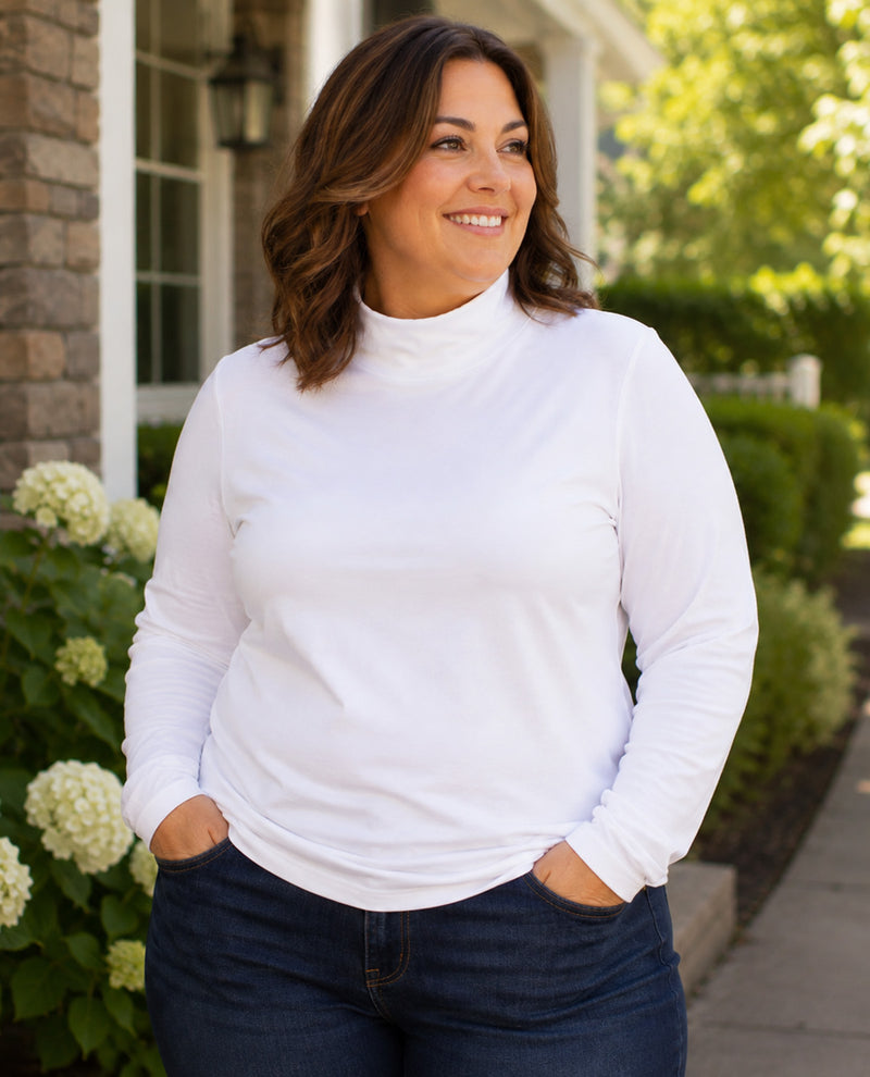 A woman wearing a white long-sleeve mock neck turtleneck top and dark blue jeans, standing outdoors in front of a house with greenery.