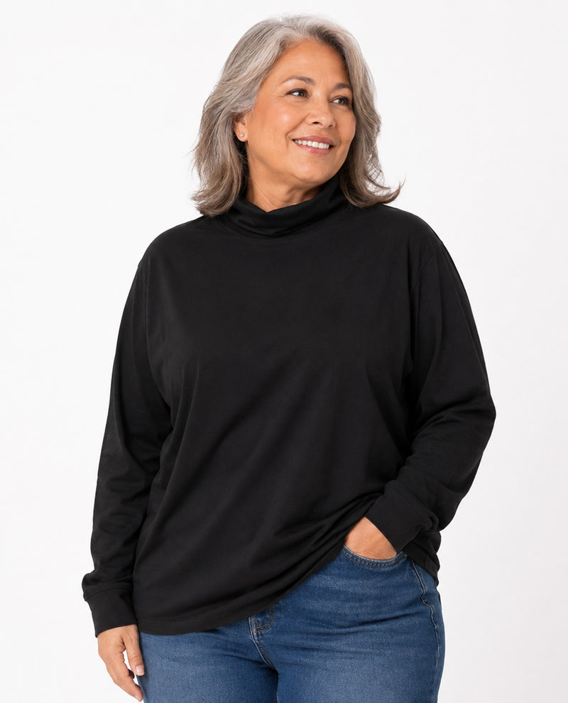 A smiling woman with gray hair wearing a black long-sleeve mock neck top and blue jeans, standing against a plain white studio background.