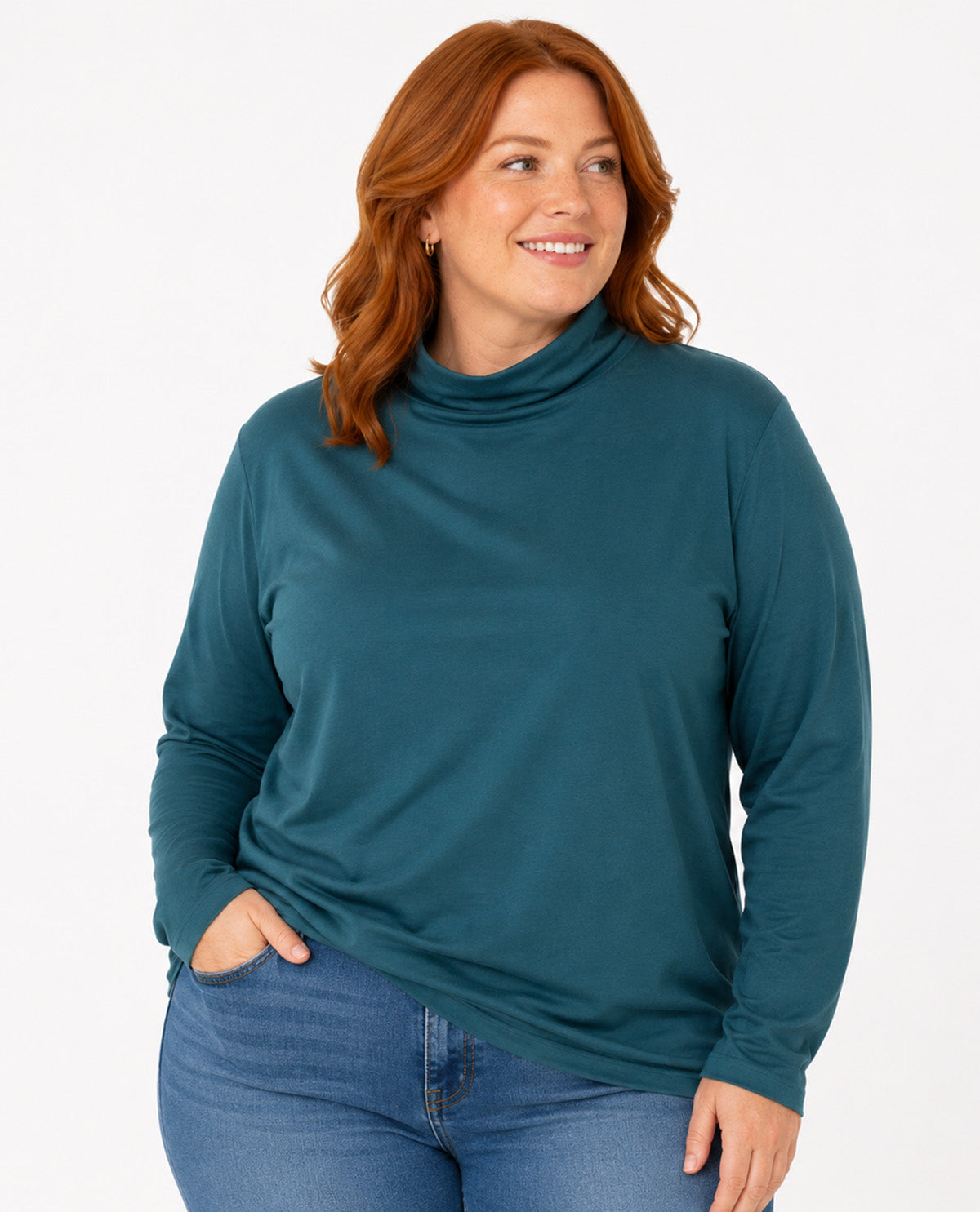 A woman with red hair wearing a dark teal long-sleeve turtleneck top and blue jeans, standing against a plain white studio background.