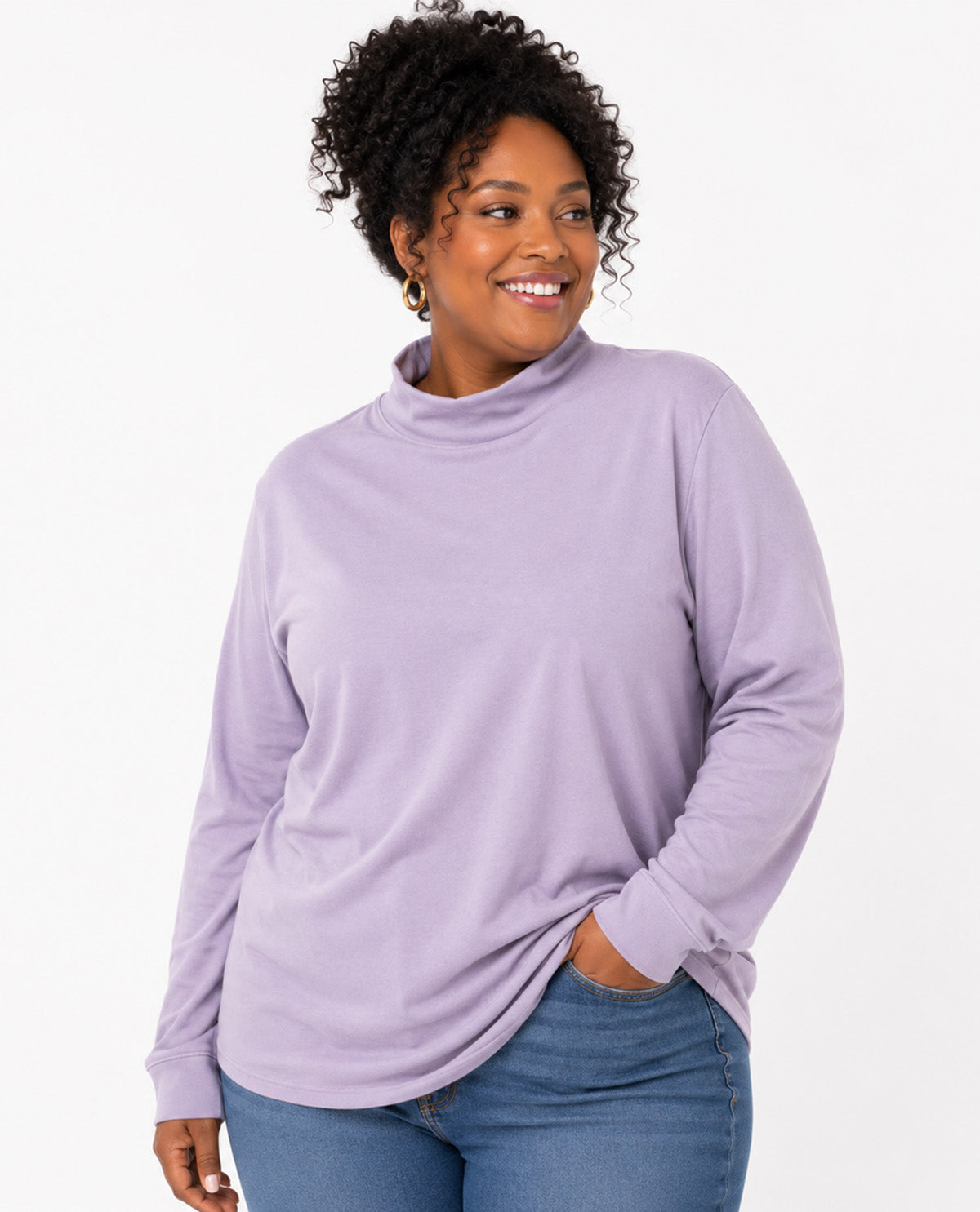 A woman with curly black hair wearing a light purple long-sleeve mock neck top and blue jeans, standing against a plain white background.