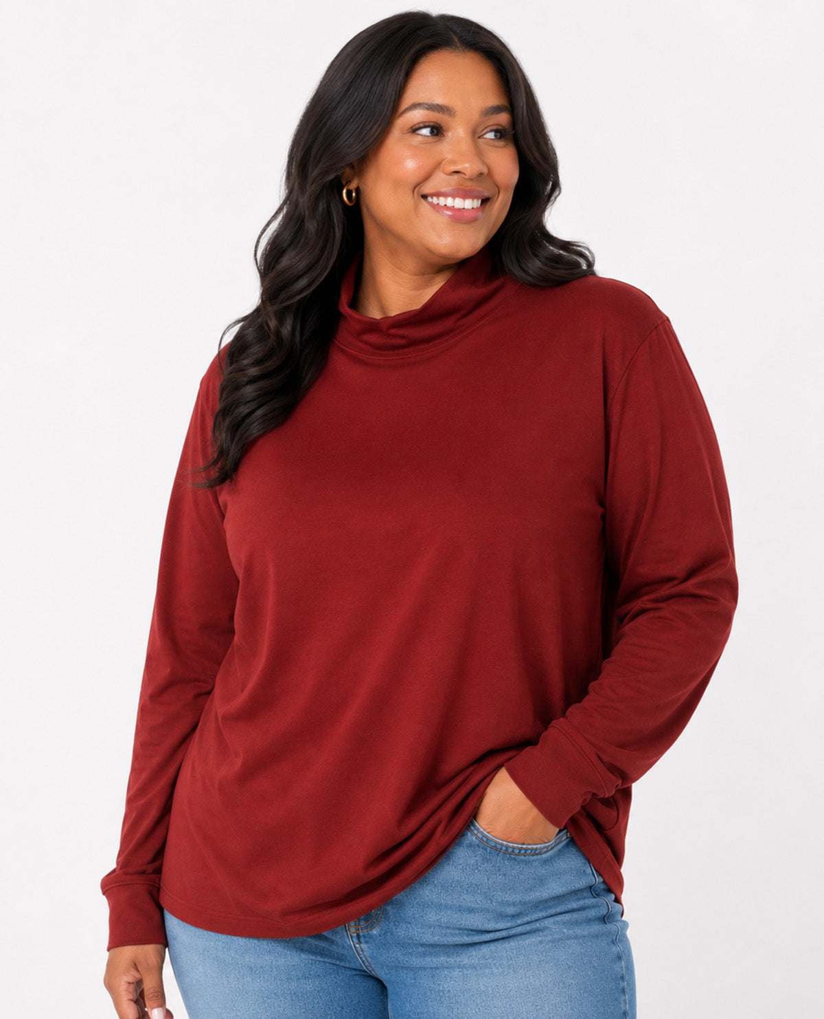 A woman wearing a long-sleeve cowl neck top in a deep burgundy red, paired with blue jeans, against a plain white studio background.