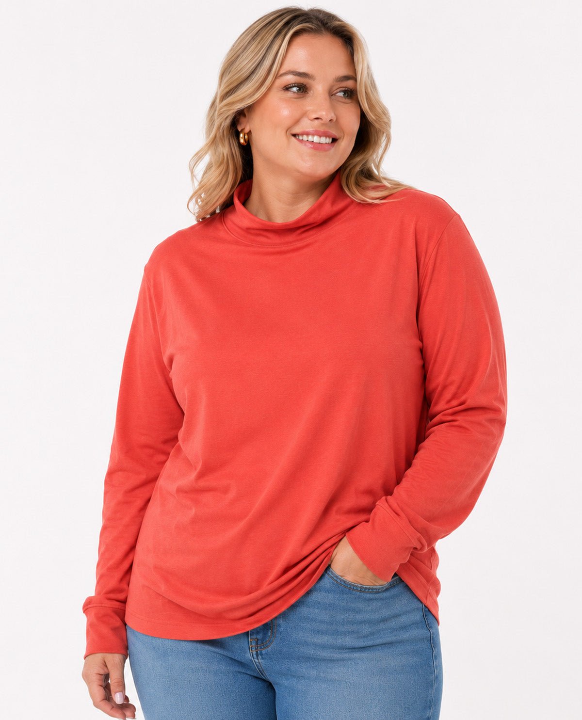 A woman wearing a coral orange long-sleeve mock neck top and blue jeans, standing against a plain white studio background.