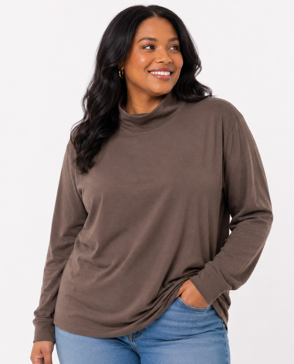 A woman wearing a brown long-sleeve mock neck top and blue jeans, posing against a solid white background.