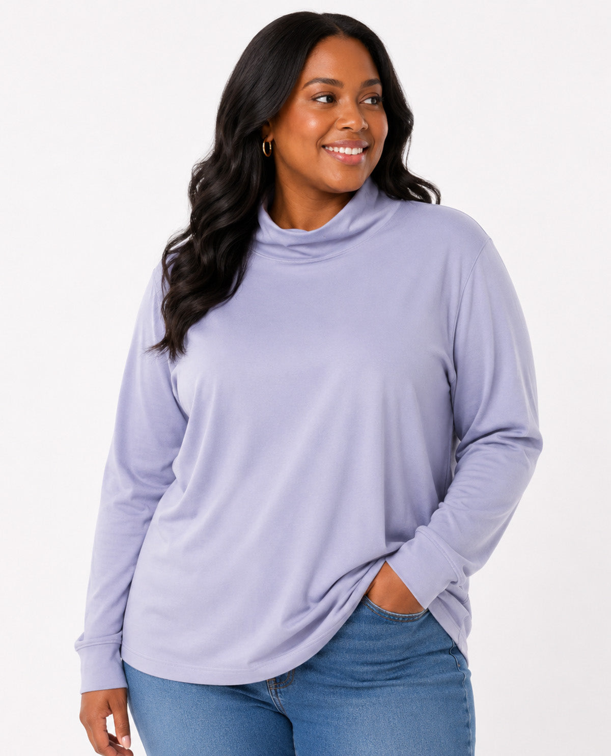 A woman wearing a light lavender long-sleeve mock neck top and blue jeans, standing against a plain white studio background.