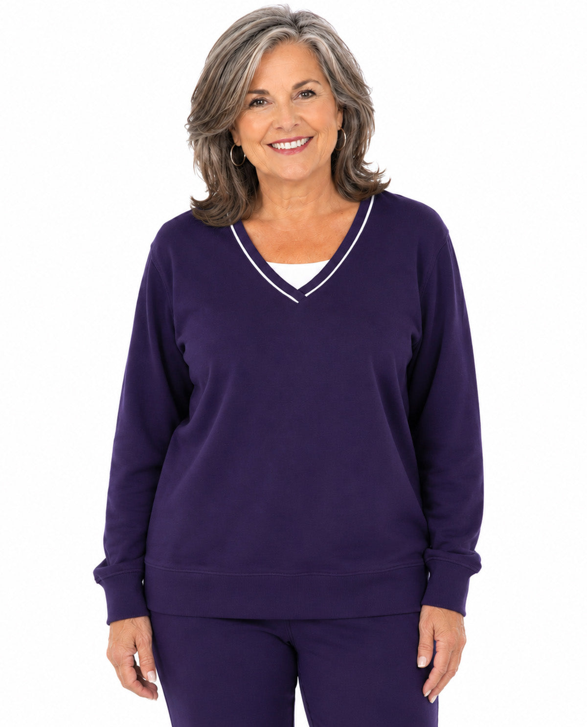 A smiling woman with shoulder-length gray hair wearing a dark purple long-sleeve V-neck sweatshirt with white trim and matching purple pants, photographed against a plain white background.