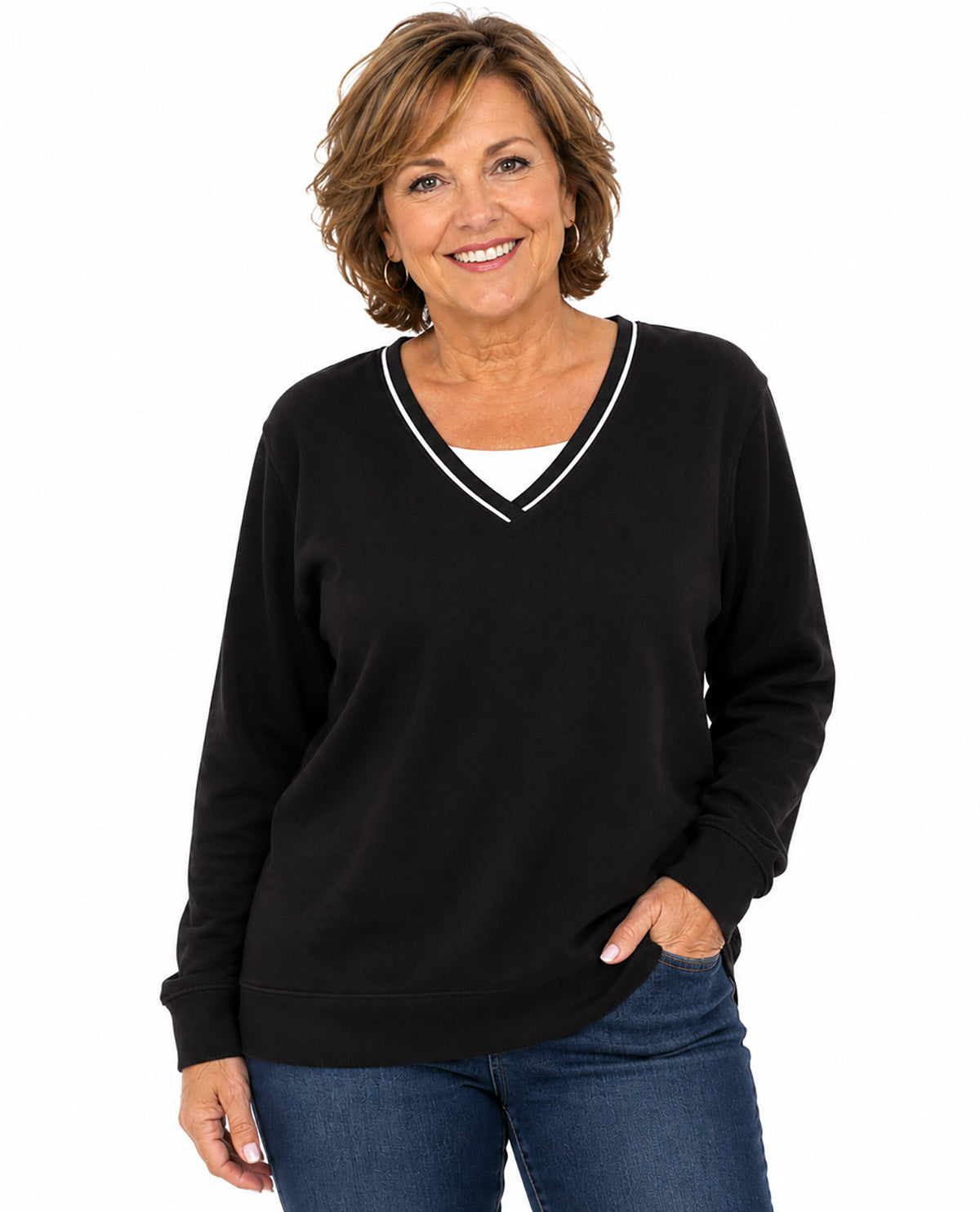 A smiling woman with short brown hair wearing a black long-sleeve V-neck sweater with white trim, paired with blue jeans, against a plain white background.