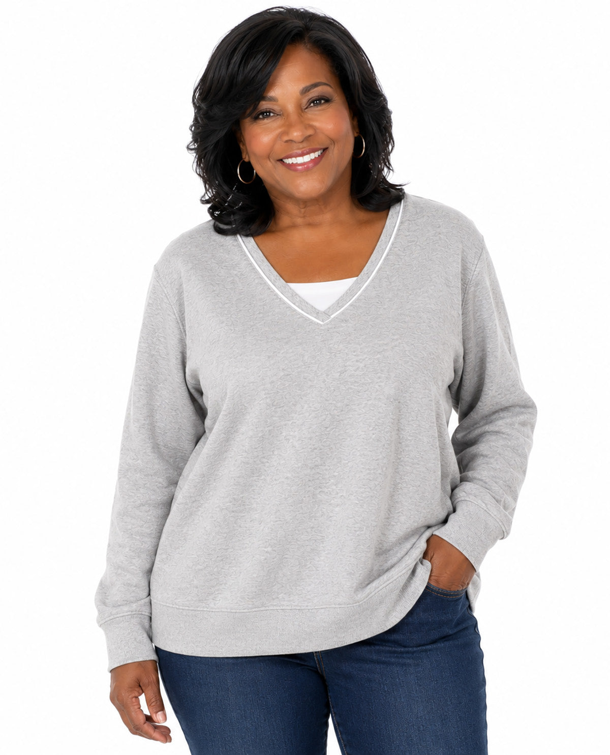 A woman wearing a light gray heather V-neck long-sleeve top with white trim, paired with dark blue jeans, against a plain white background.