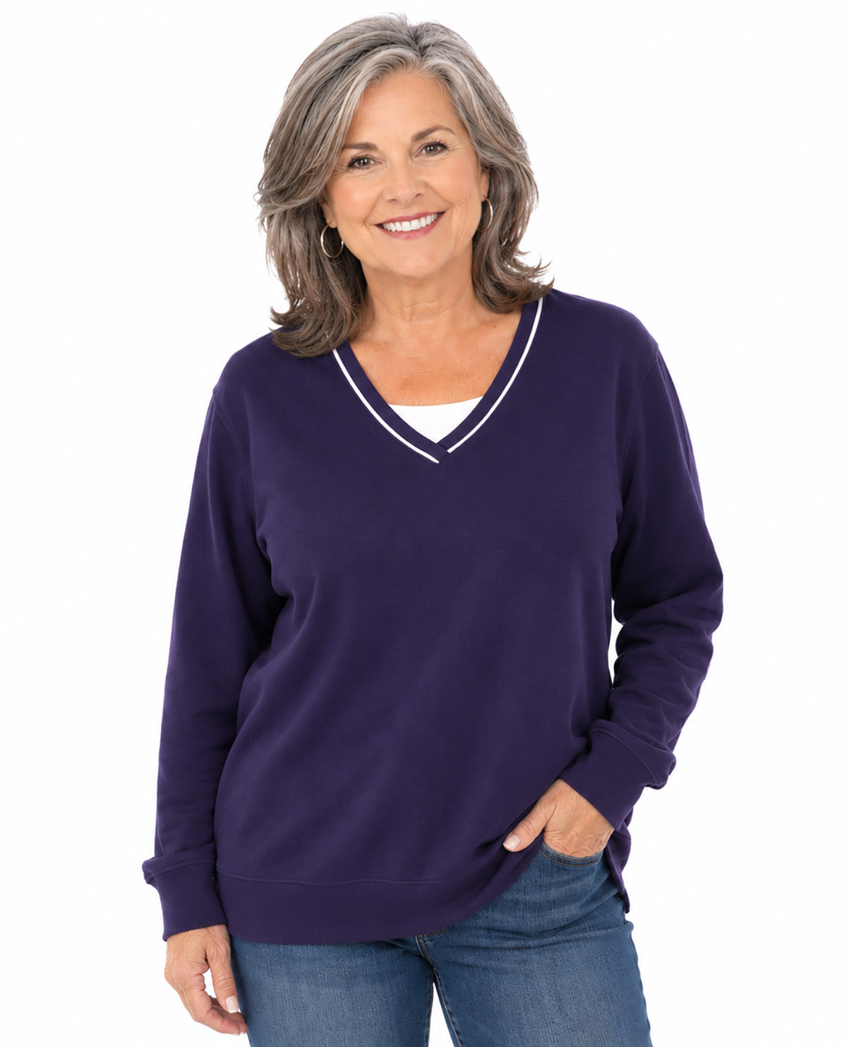 A smiling woman with shoulder-length gray hair wears a dark purple long-sleeve V-neck sweater with thin white trim, paired with blue jeans, against a plain white background.