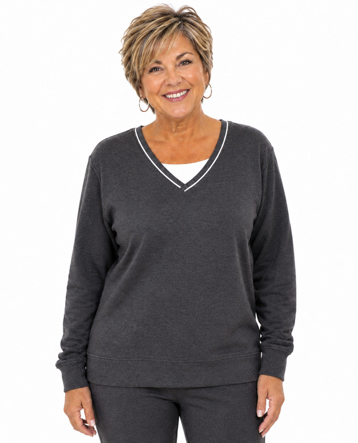 A smiling woman with short blonde hair wearing a dark gray long-sleeve V-neck sweater with white trim over a white undershirt, paired with matching dark gray pants, against a plain white background.
