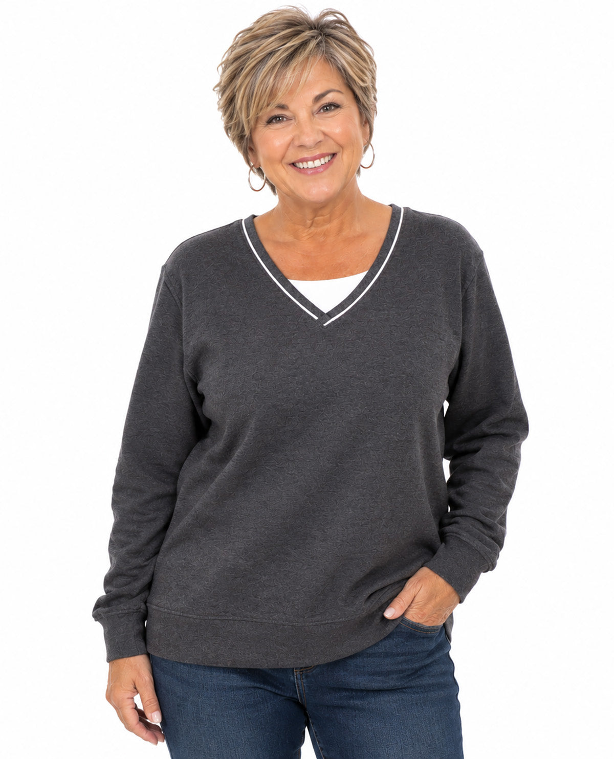 A smiling woman with short blonde hair wearing a dark gray long-sleeve V-neck sweater with white trim, layered over a white undershirt and paired with blue jeans, against a plain white background.