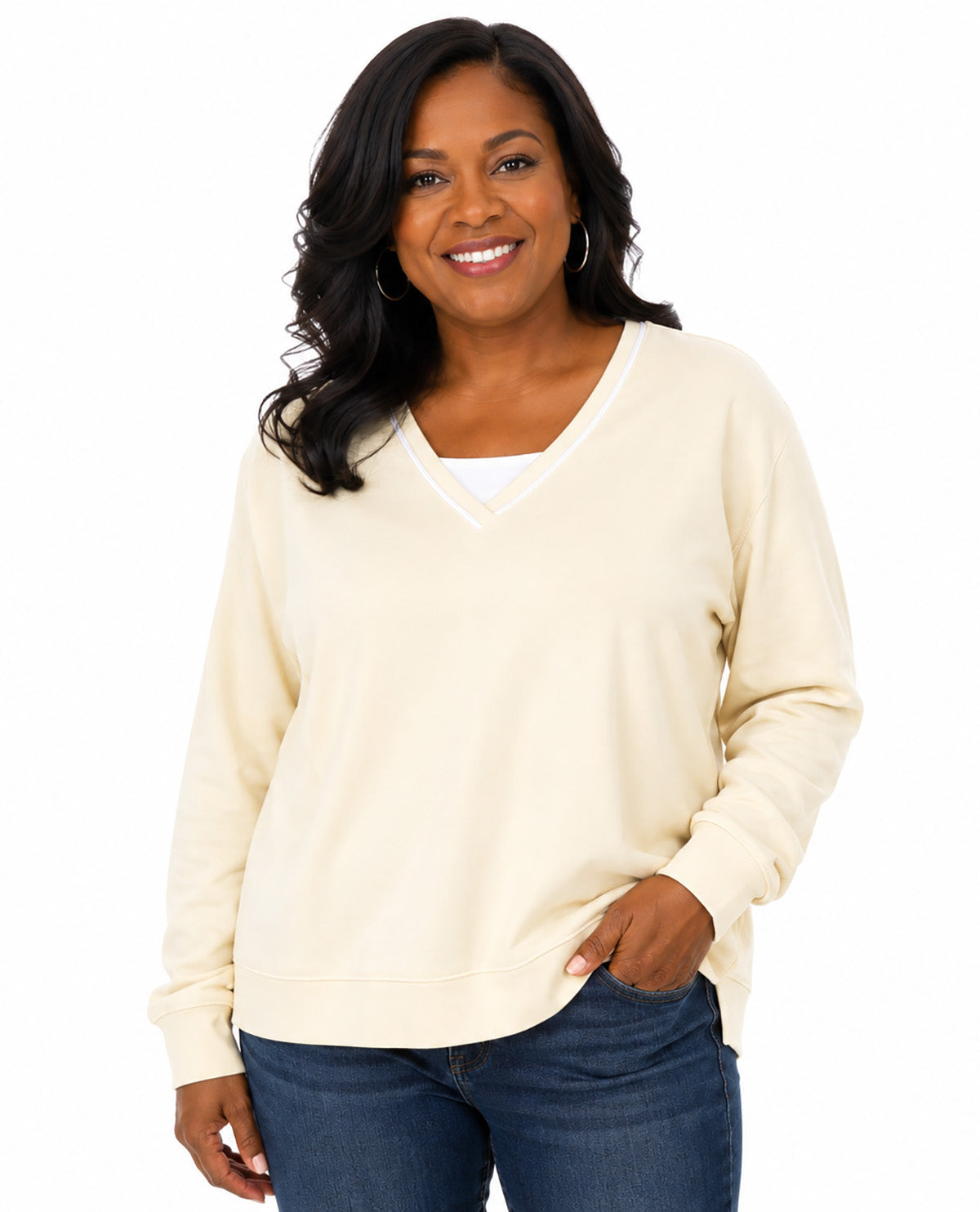 A smiling woman with long dark hair wearing a cream-colored long-sleeve V-neck sweater over a white undershirt and blue jeans, photographed against a solid white background.