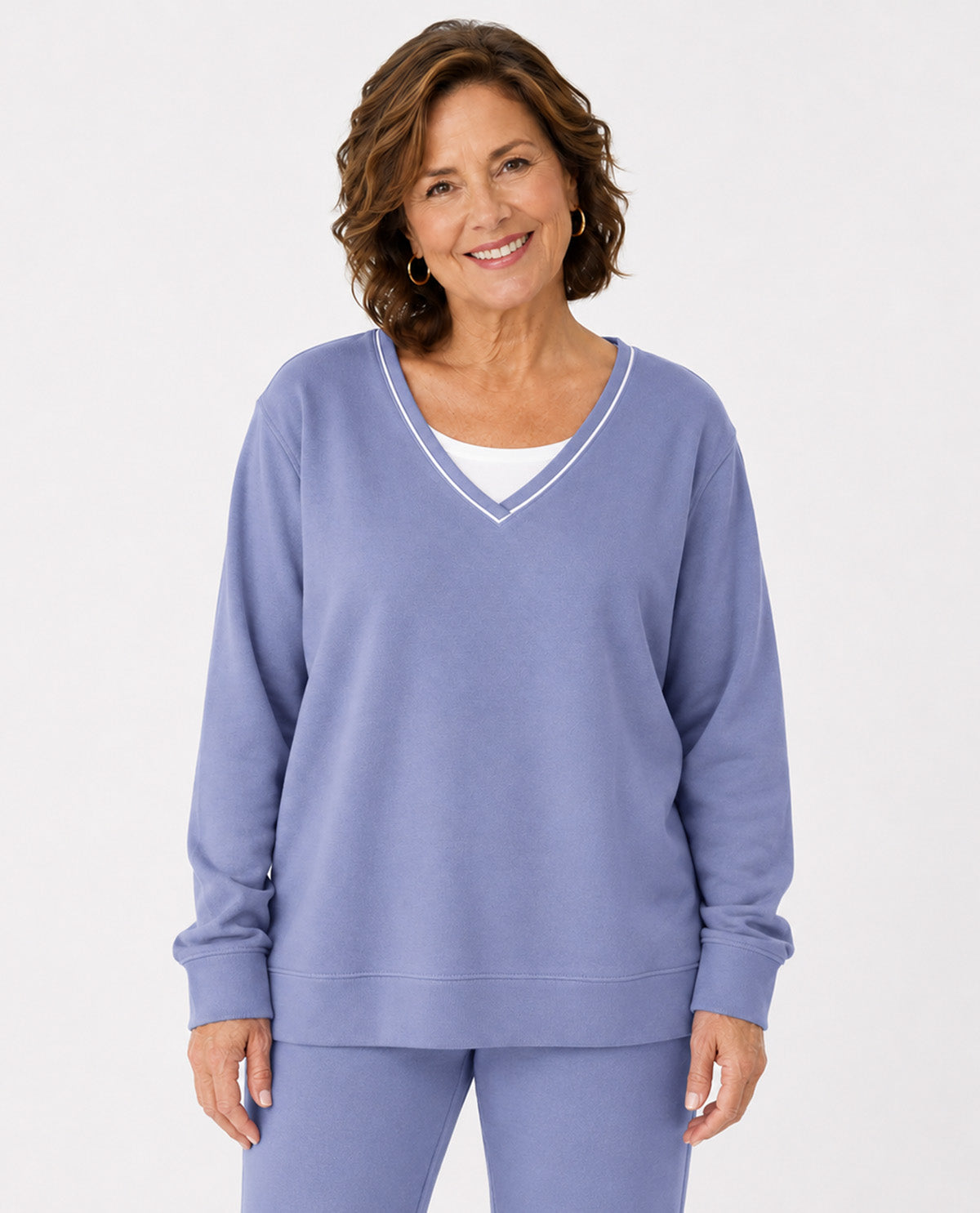 A smiling woman wearing a lavender-purple V-neck sweatshirt with white trim over a white undershirt, paired with matching purple sweatpants, against a light gray studio background.