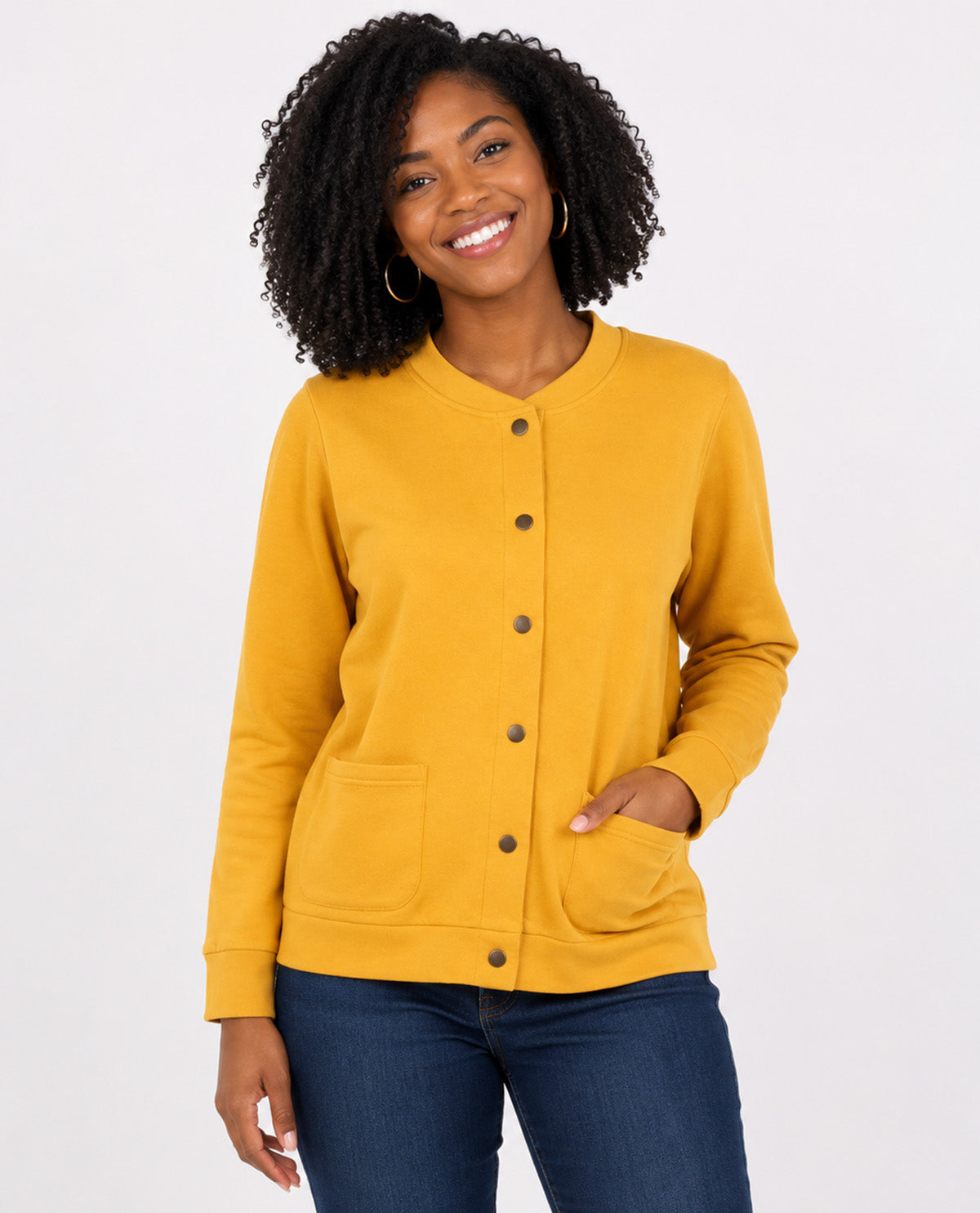 A woman wearing a mustard yellow button-up cardigan sweater with front pockets, paired with dark blue jeans, against a plain light gray studio background.