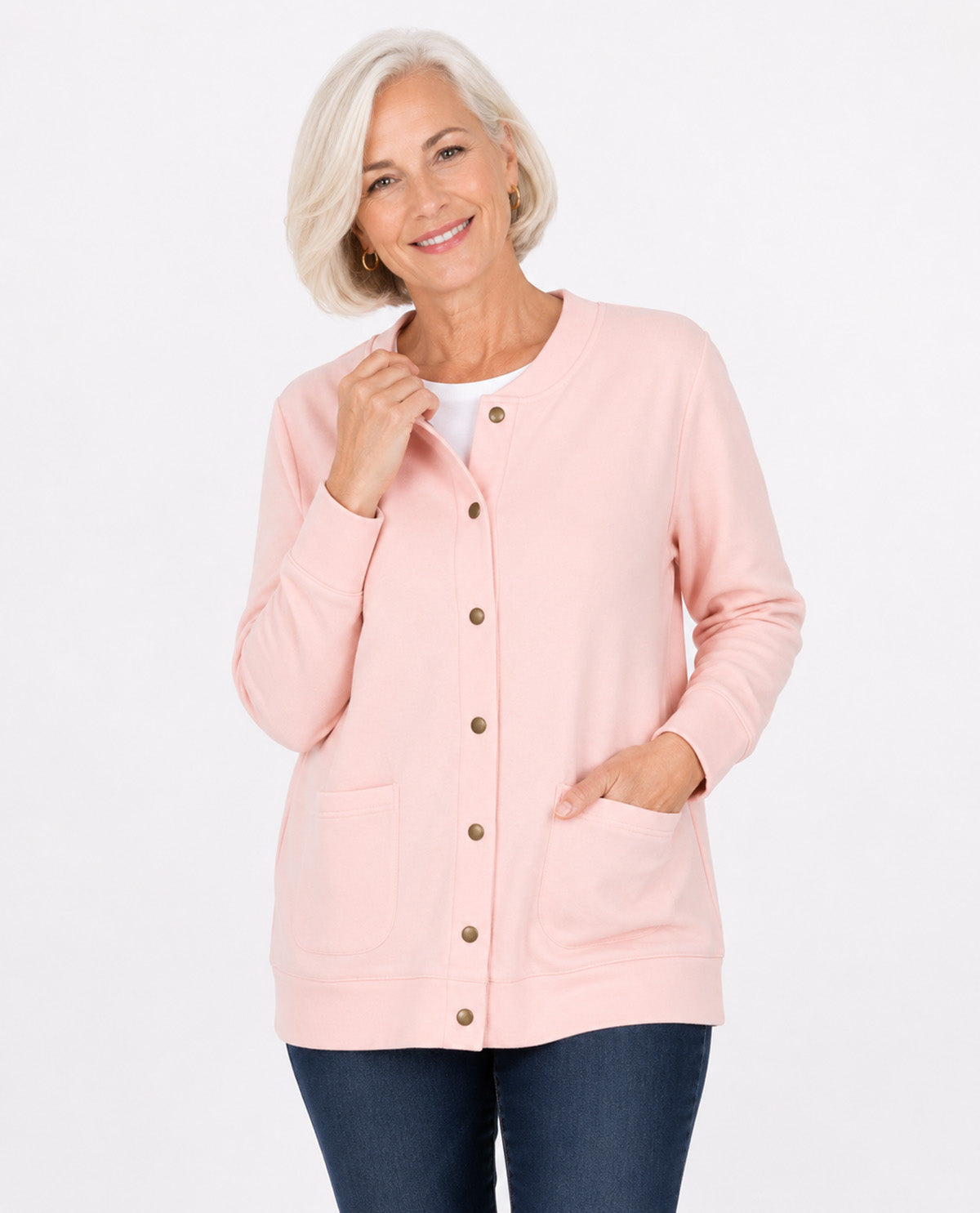 A smiling older woman with short blonde hair models a light pink button-front cardigan with front pockets over a white top and dark blue jeans, shot against a plain white studio background.
