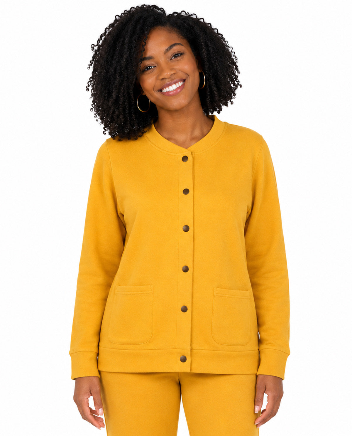 A smiling woman with curly black hair wearing a mustard yellow button-front cardigan and matching pants, photographed against a plain white studio background.
