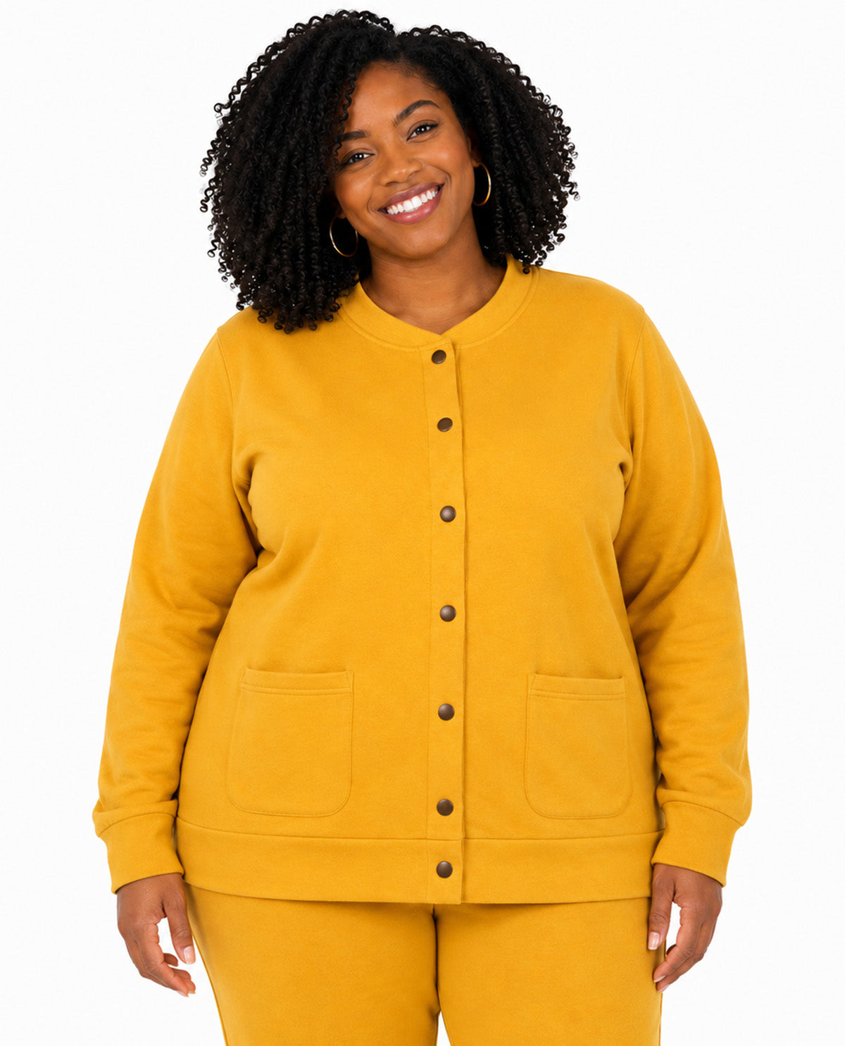 A smiling woman with curly black hair wearing a mustard yellow button-front cardigan and matching pants against a plain white studio background.