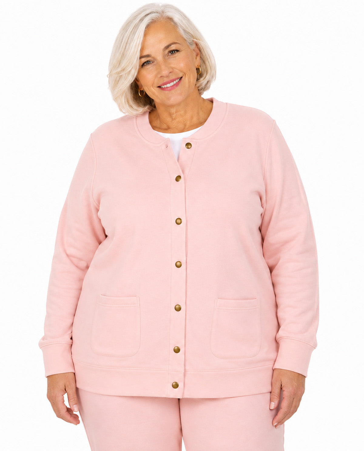 A smiling older woman with short blonde hair wears a light pink button-front cardigan with two front patch pockets and matching pink pants against a plain white studio background.