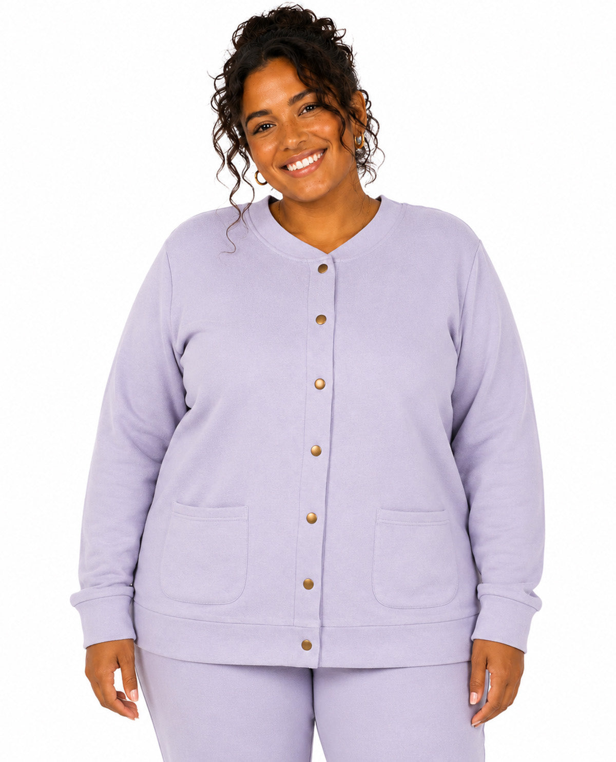 A smiling woman wearing a light lavender cardigan with gold buttons and matching lavender pants, photographed against a plain white studio background.