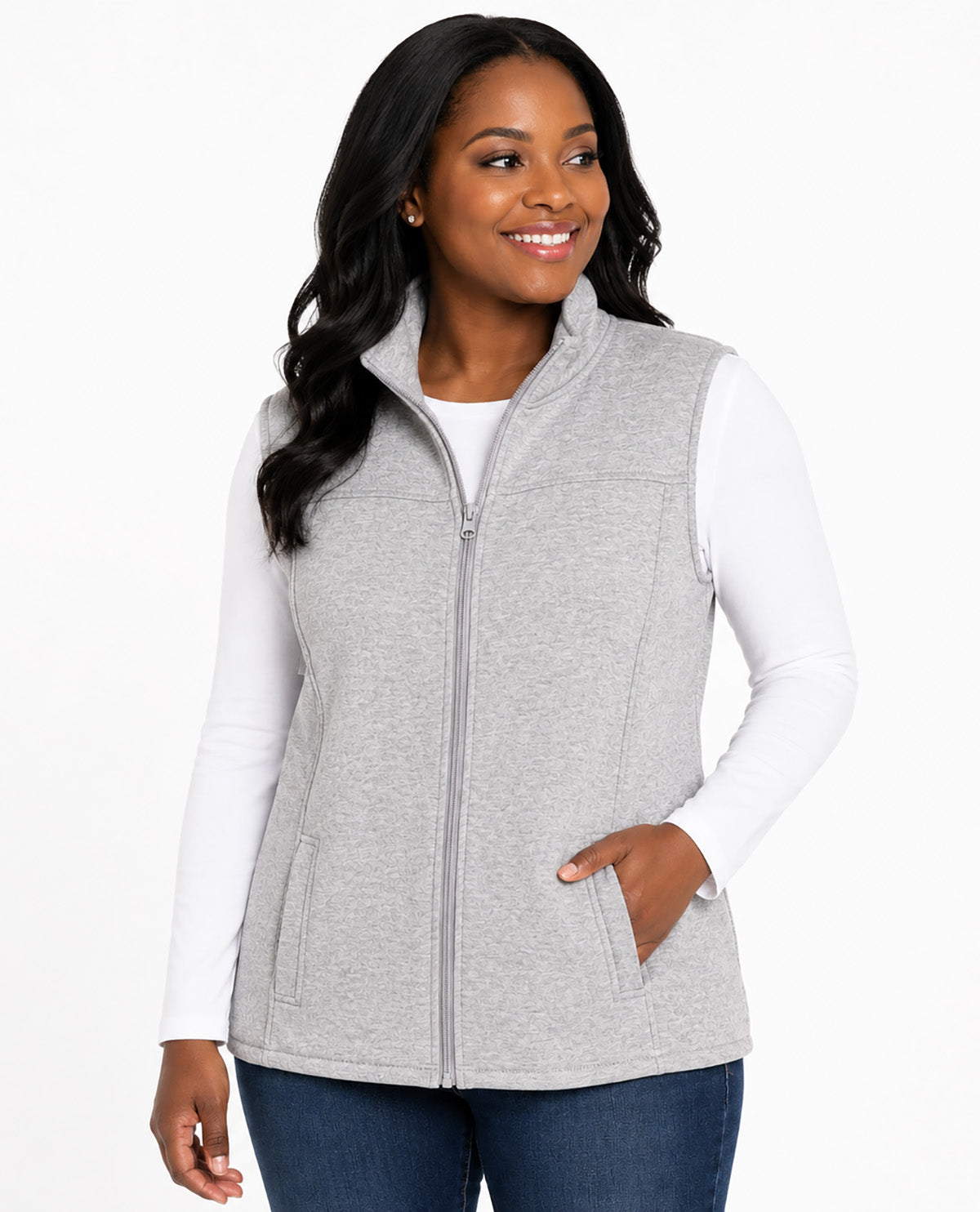 A woman wearing a light gray zip-up fleece vest over a white long-sleeve shirt and blue jeans, photographed against a clean white studio background.