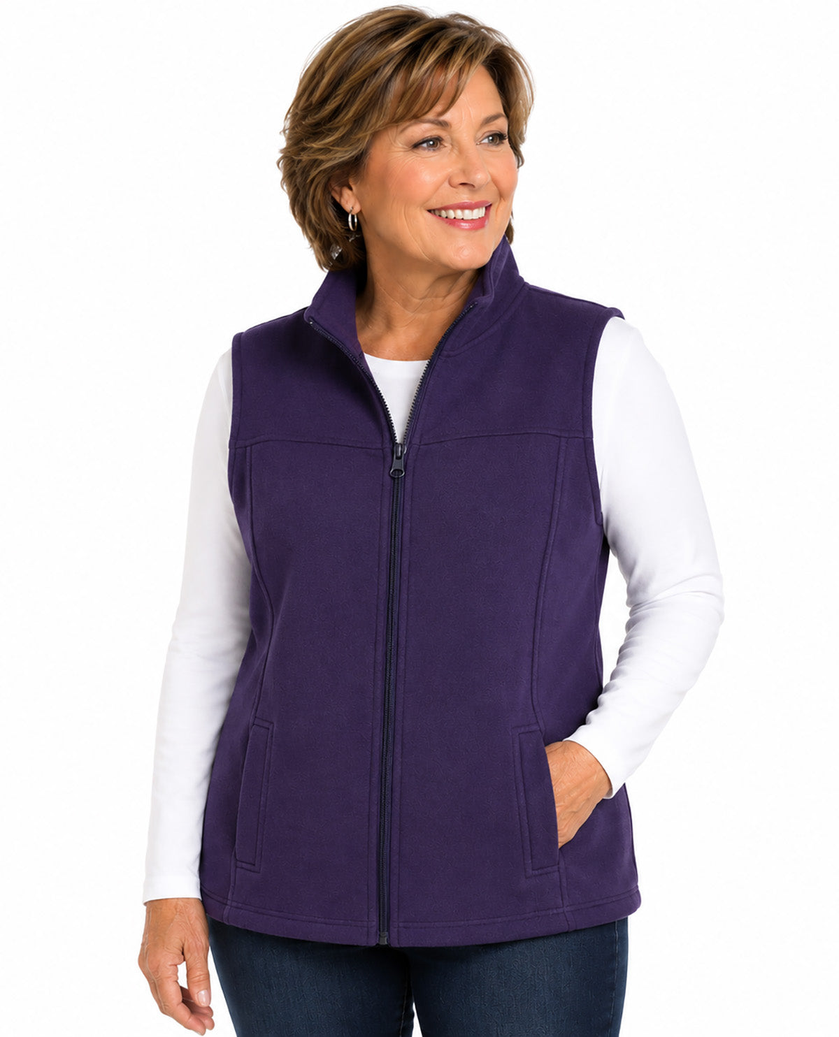A smiling woman wearing a purple zip-up fleece vest over a white long-sleeve shirt and dark blue jeans, photographed against a plain white studio background.
