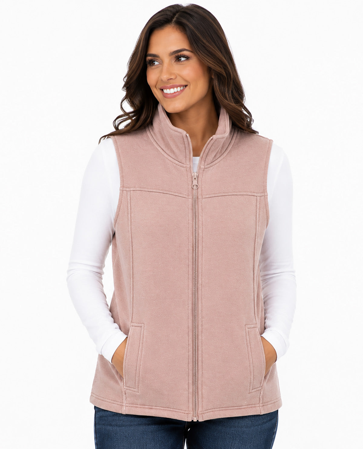 A woman wearing a dusty pink zip-up fleece vest over a white long-sleeve shirt and blue jeans, standing against a plain white background.