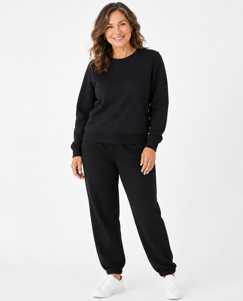 A woman modeling a black crewneck sweatshirt and matching black jogger pants, paired with white sneakers, against a plain white studio background.