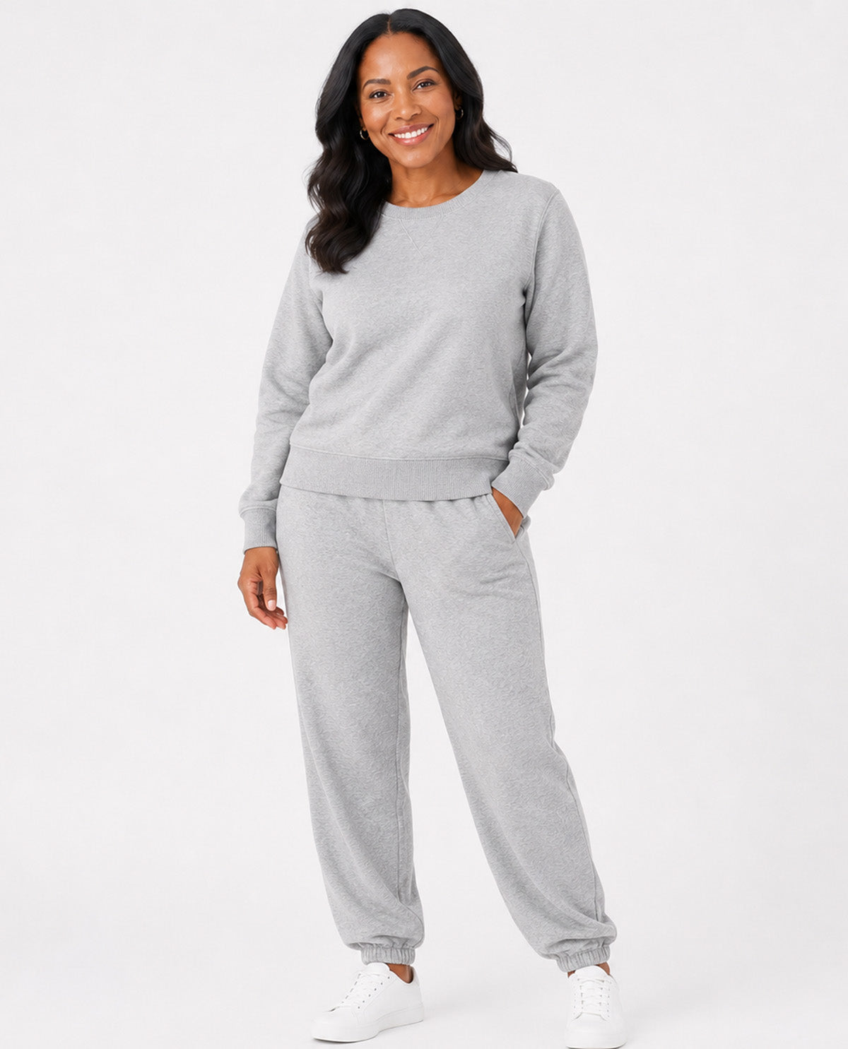 A woman modeling a light gray heather sweatshirt and matching jogger pants set, paired with white sneakers, against a plain white studio background.