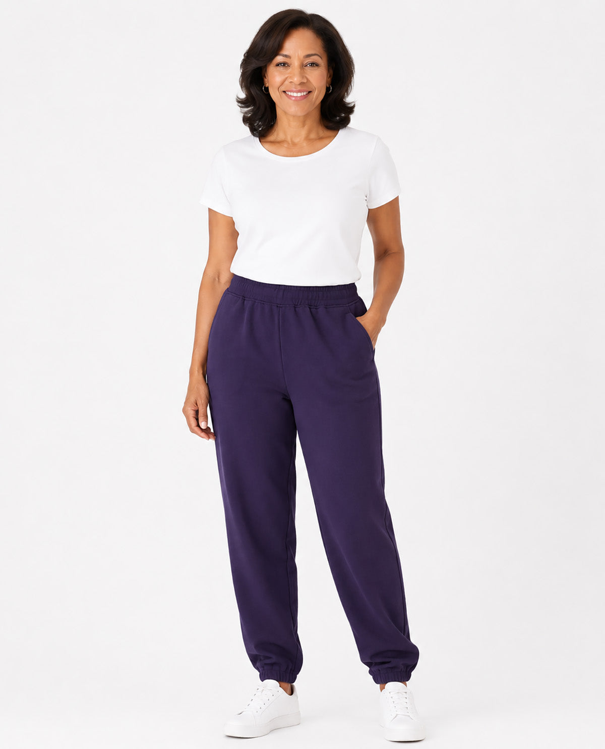 A woman modeling dark purple sweatpants with a white t-shirt and white sneakers, standing against a solid white studio background.
