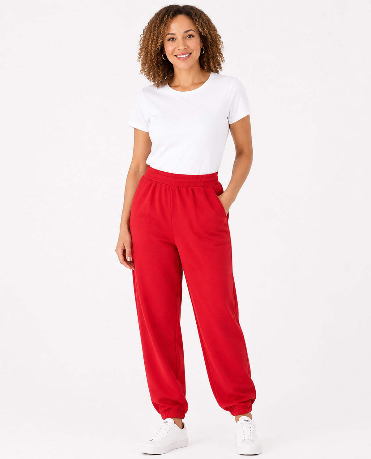 A woman modeling bright red jogger sweatpants with a white t-shirt and white sneakers against a plain white studio background.