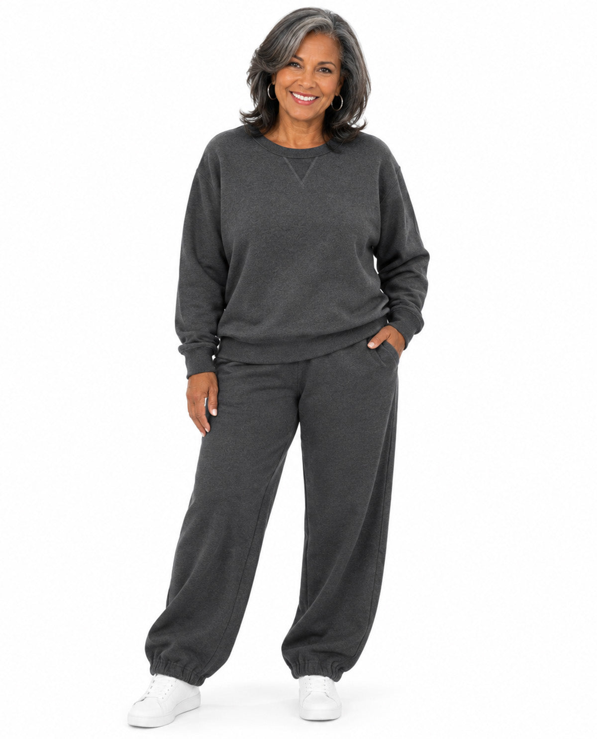 A smiling woman with gray hair is modeling a dark gray two-piece loungewear set, consisting of a long-sleeve crewneck sweatshirt and matching jogger pants, paired with white sneakers against a plain white background.