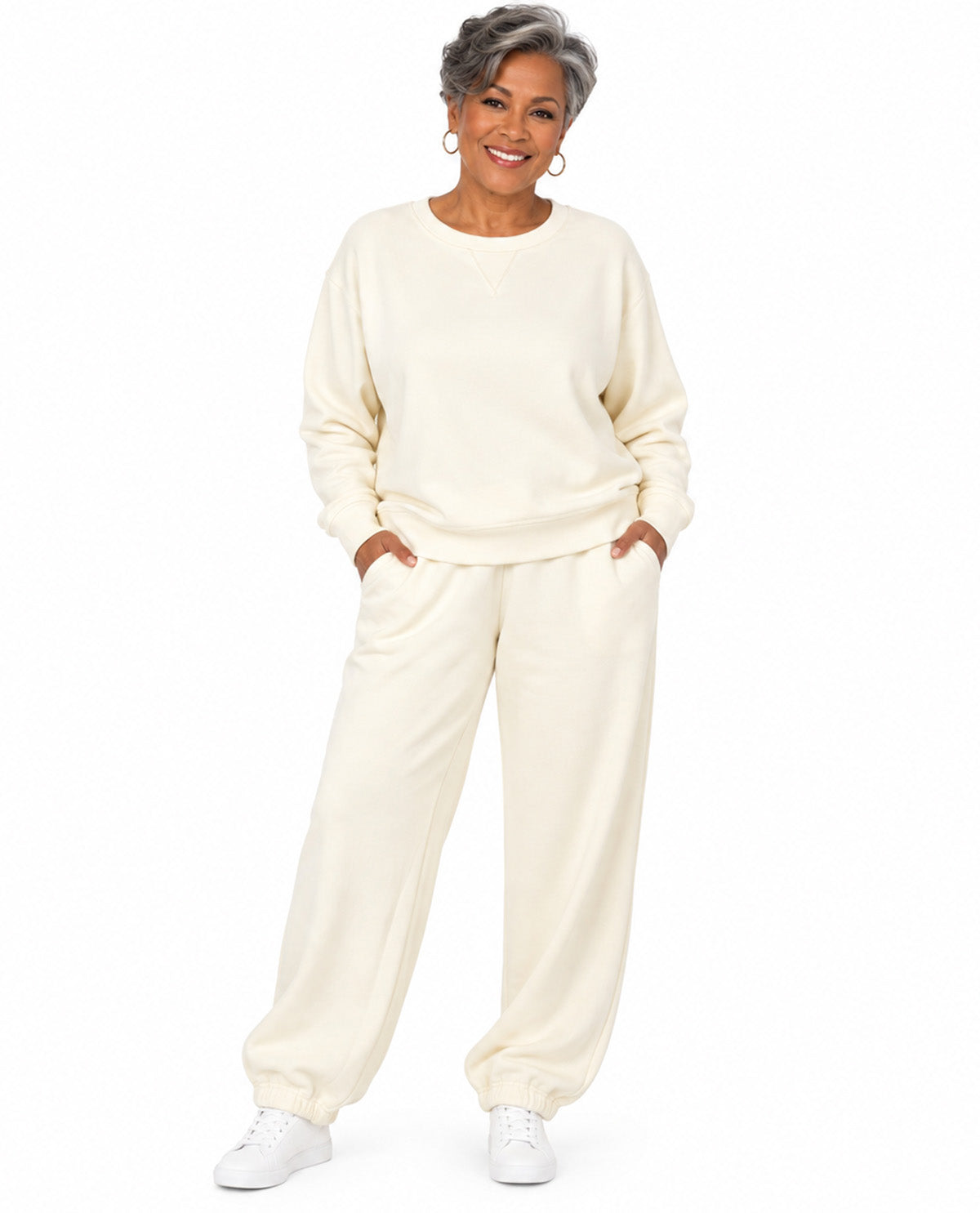 A smiling woman with short gray hair models a matching cream-colored lounge set consisting of a long-sleeve sweatshirt and wide-leg sweatpants, paired with white sneakers against a solid white background.