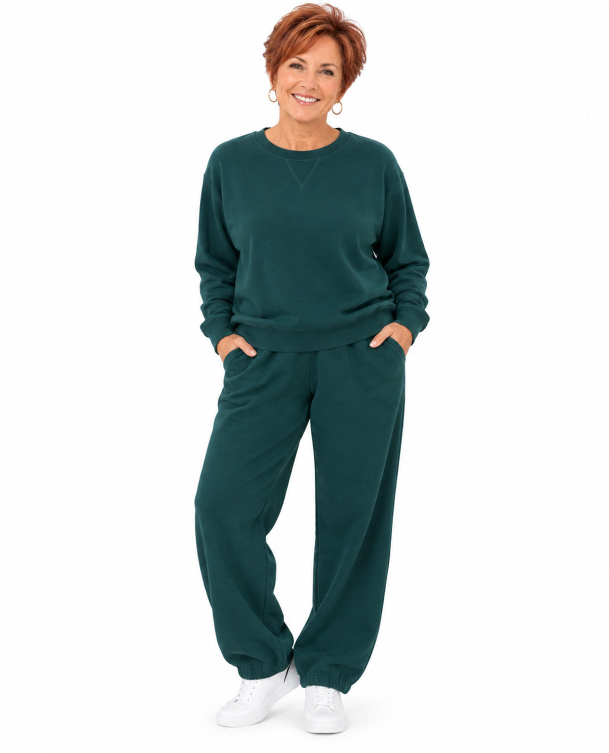 A smiling woman with short red hair stands against a plain white background wearing a matching dark green sweatshirt and sweatpants set, with her hands in the pockets of the pants and white sneakers.