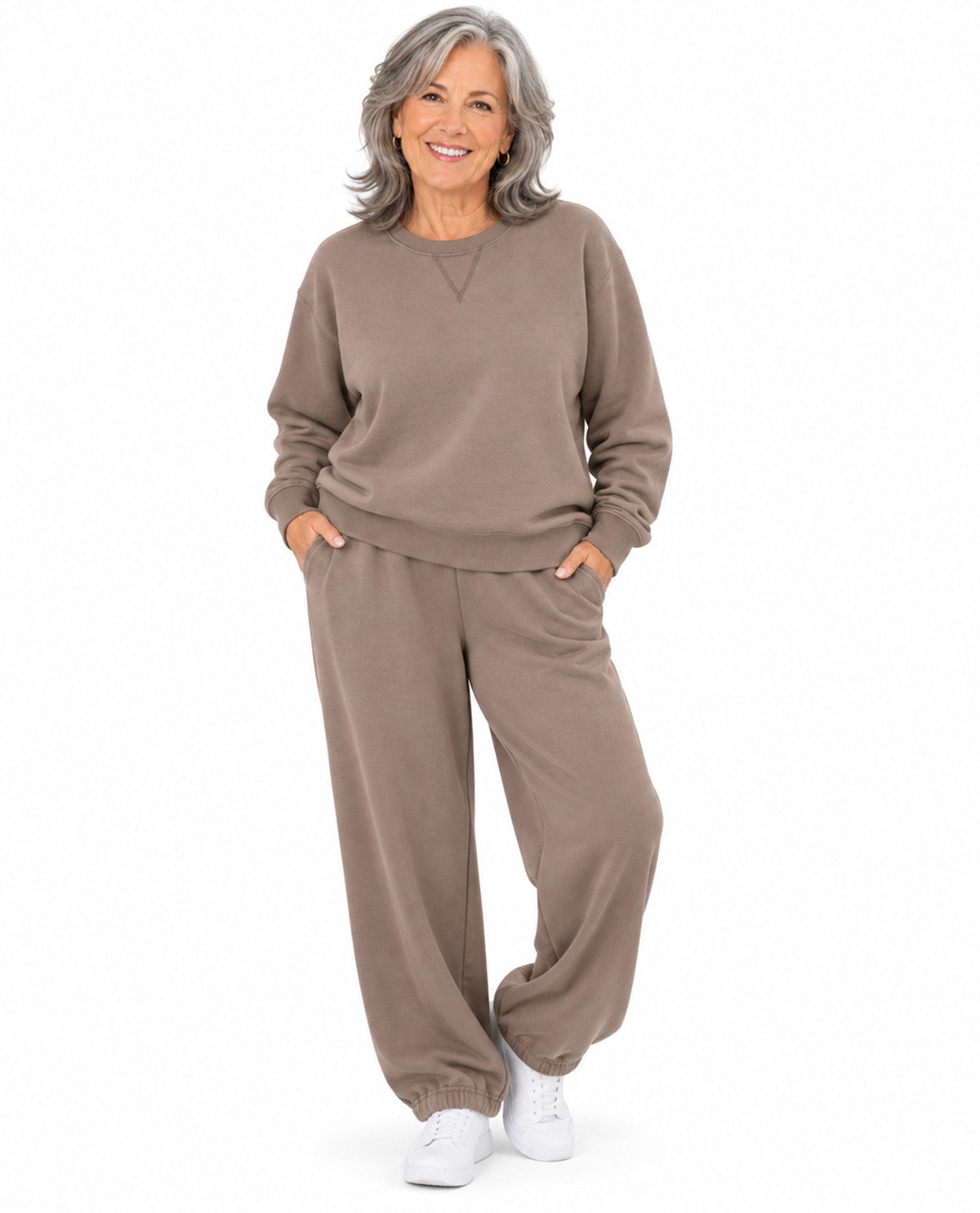 A smiling older woman stands against a plain white studio background wearing a matching taupe-brown lounge set consisting of a long-sleeve crewneck sweatshirt and wide-leg jogger pants, paired with white sneakers.