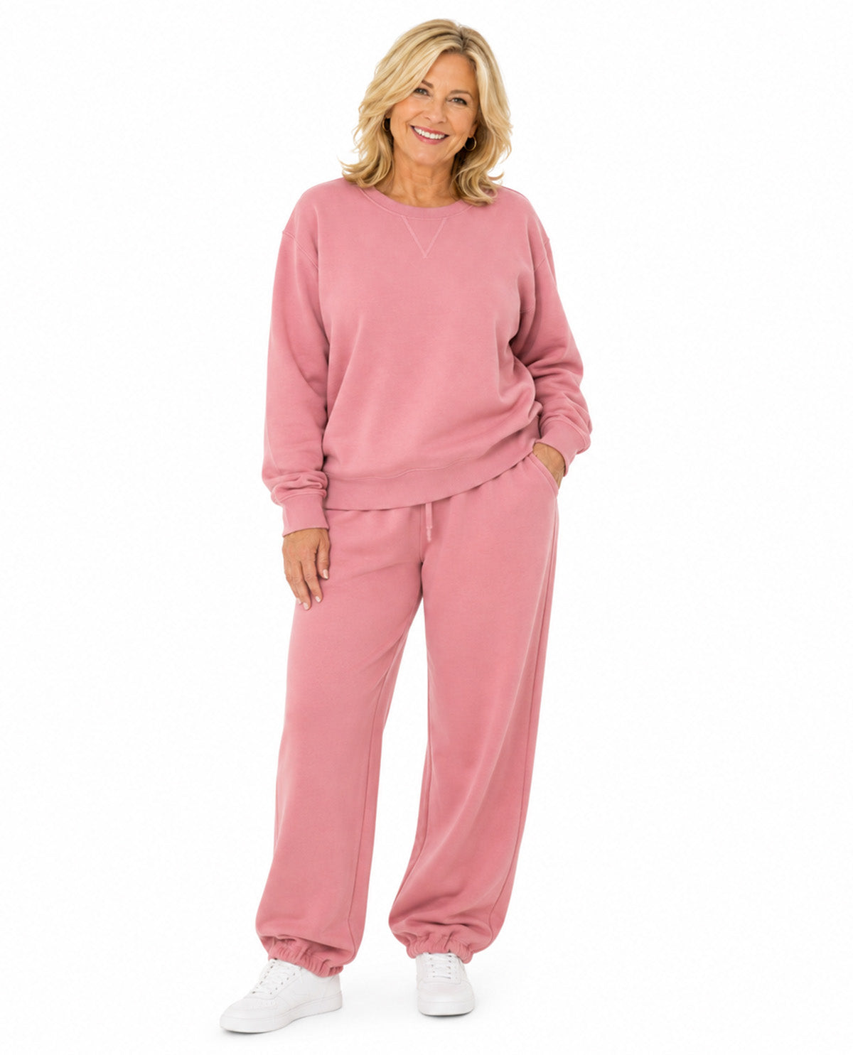 A smiling blonde woman modeling a matching dusty pink lounge set consisting of a long-sleeve sweatshirt and jogger pants, paired with white sneakers against a plain white background.