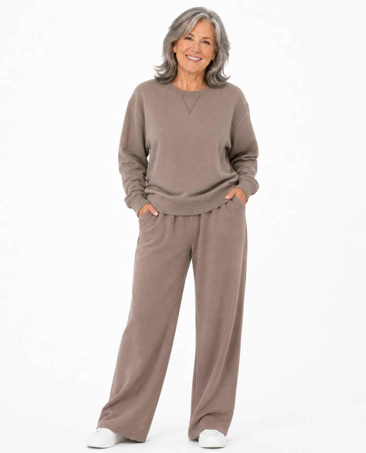 A smiling woman with gray hair stands against a plain white background wearing a matching taupe-brown lounge set consisting of a long-sleeve crewneck sweatshirt and wide-leg pants, paired with white sneakers.