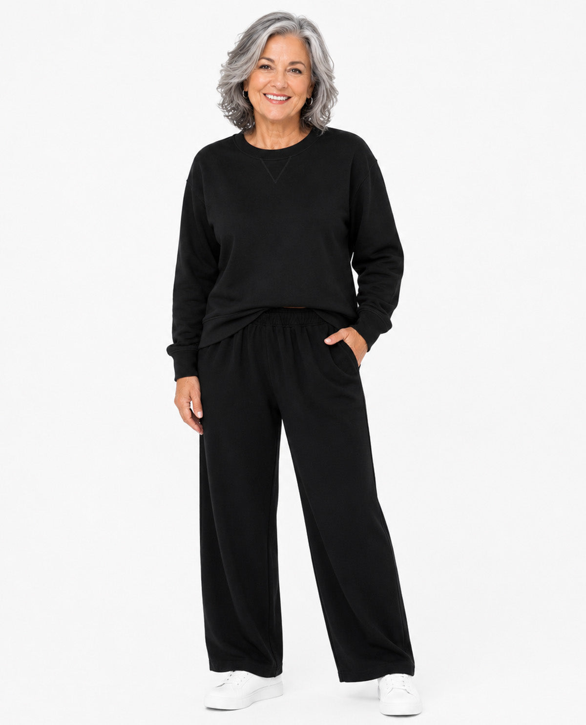 A smiling woman with gray hair stands against a plain white studio background wearing a black long-sleeve top and matching black wide-leg pants, with white sneakers.