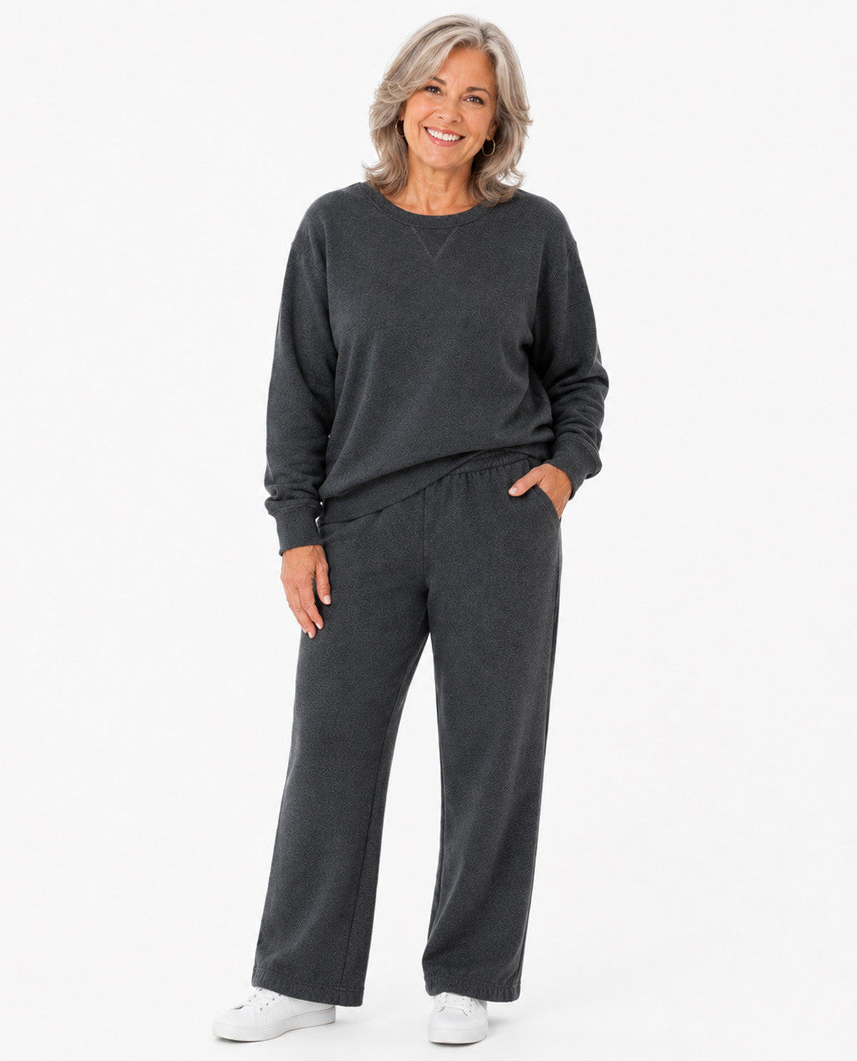 A smiling older woman stands against a clean white studio background wearing a dark charcoal gray lounge set consisting of a long-sleeve crewneck sweatshirt and matching wide-leg sweatpants, paired with white sneakers.