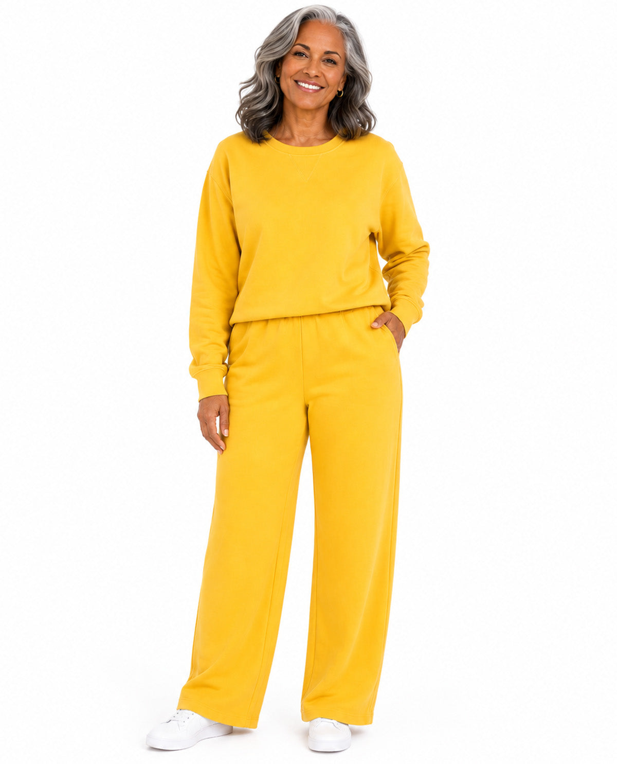 A smiling older woman with gray hair stands against a plain white background wearing a matching two-piece lounge set in bright yellow, consisting of a long-sleeve top and wide-leg pants, paired with white sneakers.