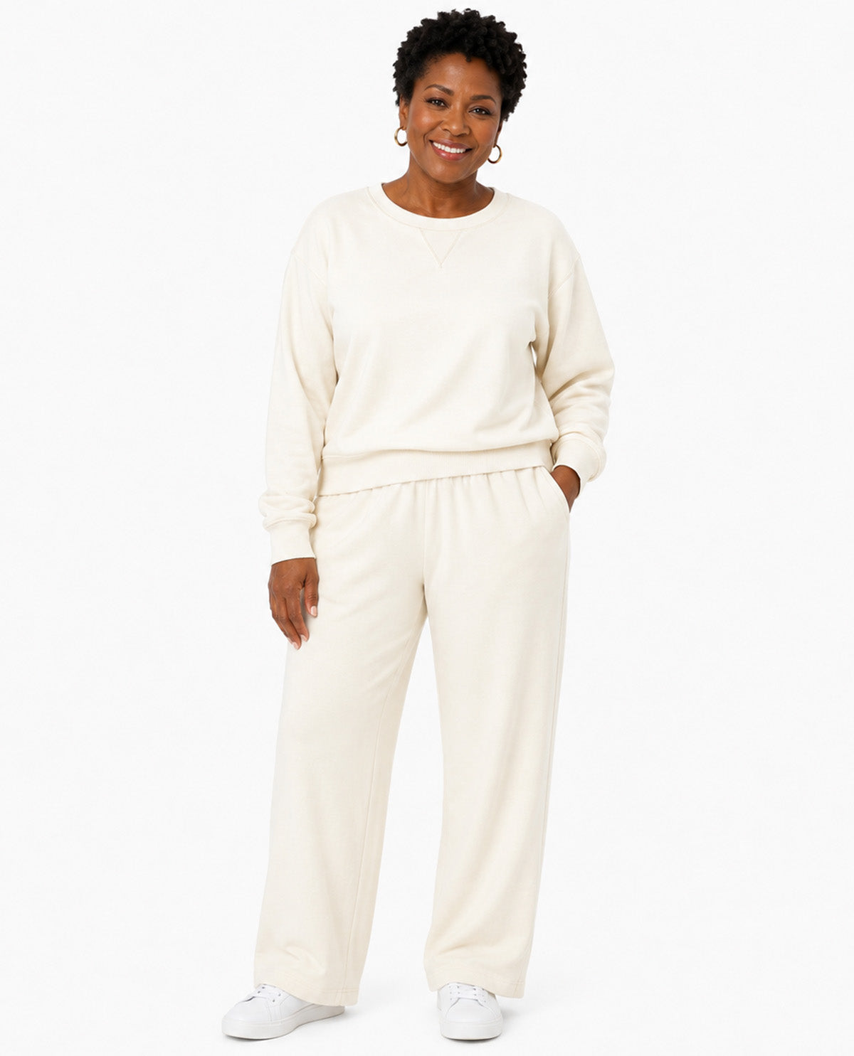 A woman modeling a matching cream-colored lounge set consisting of a long-sleeve sweatshirt and wide-leg sweatpants, paired with white sneakers against a plain white background.