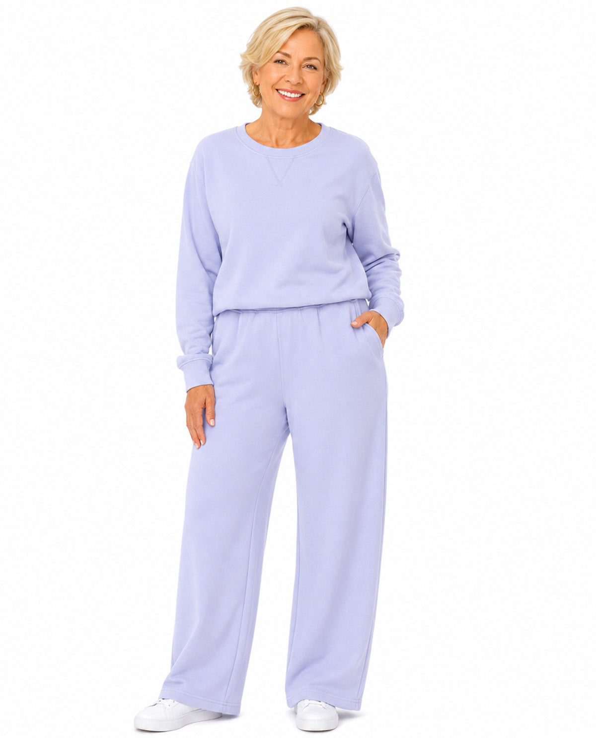 A smiling older woman modeling a light lavender two-piece loungewear set consisting of a long-sleeve crewneck sweatshirt and matching wide-leg sweatpants, paired with white sneakers against a solid white background.