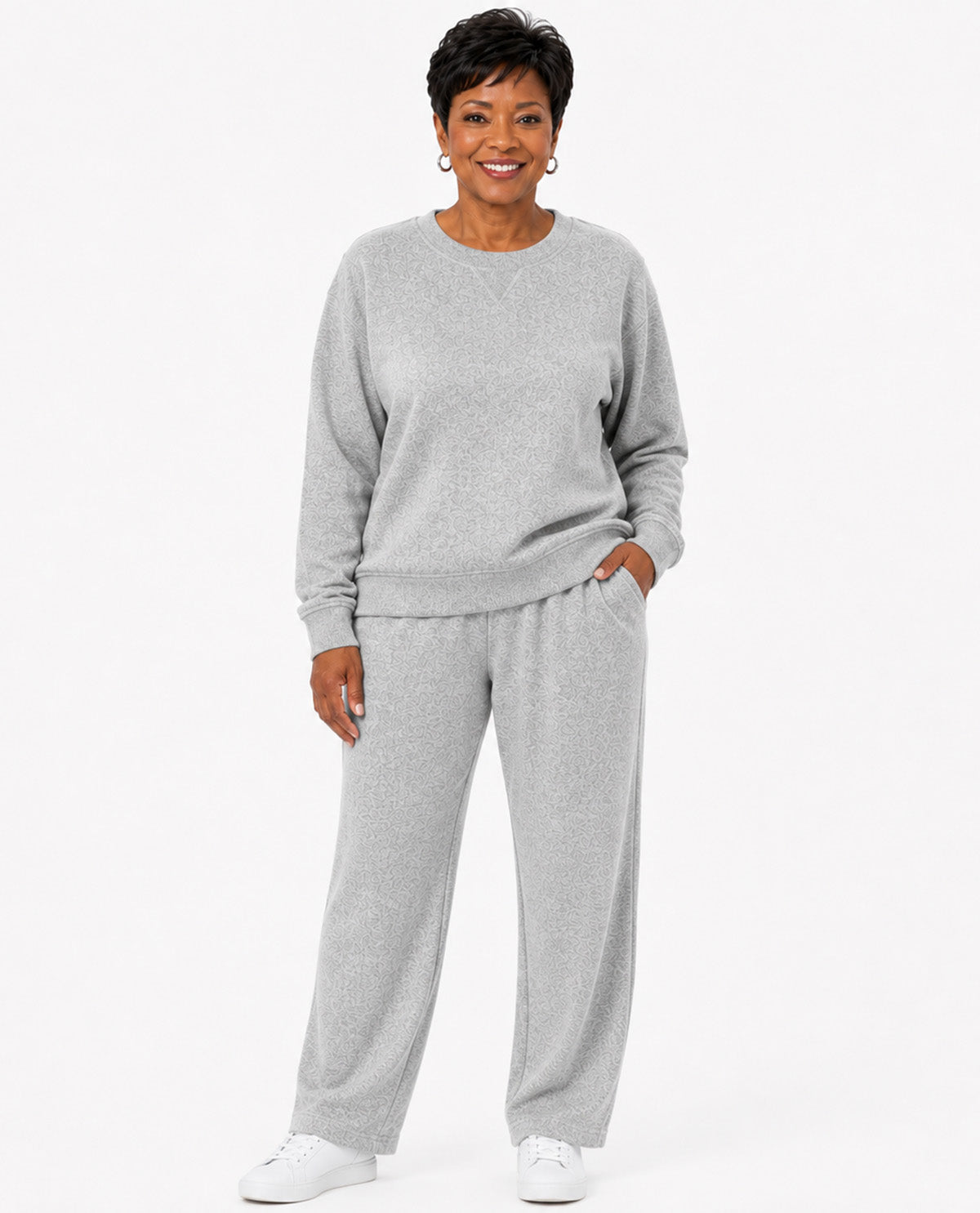 A woman modeling a light gray heathered lounge set consisting of a crewneck sweatshirt and matching wide-leg sweatpants, paired with white sneakers against a plain white studio background.