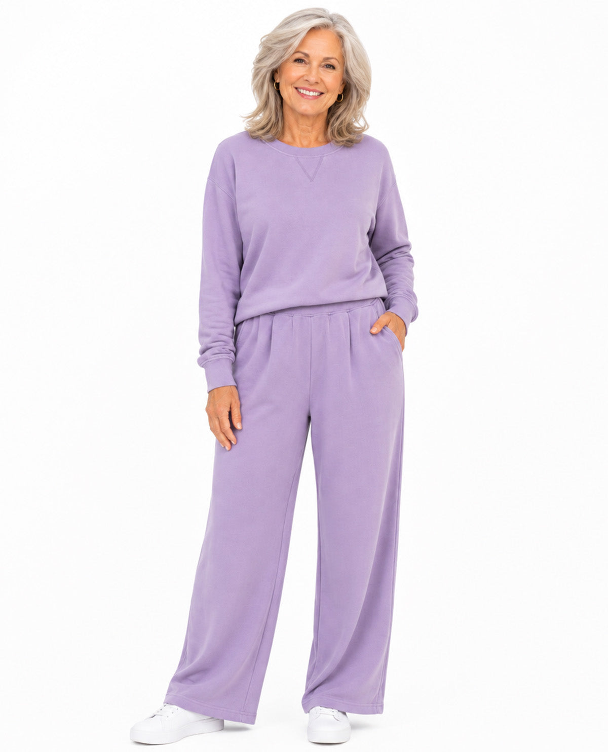 A smiling older woman stands against a plain white studio background wearing a matching lavender lounge set with a long-sleeve top and wide-leg pants, paired with white sneakers.