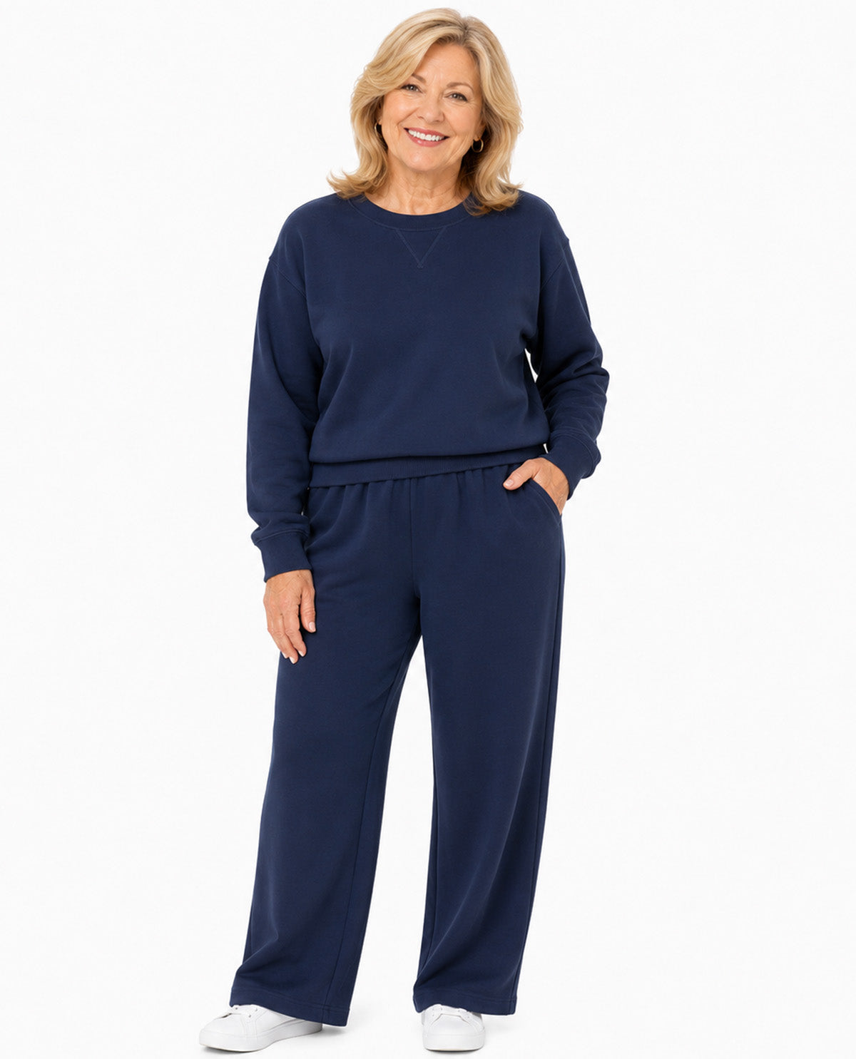 A smiling blonde woman wearing a matching navy blue sweatshirt and wide-leg sweatpants set, paired with white sneakers, against a plain white studio background.