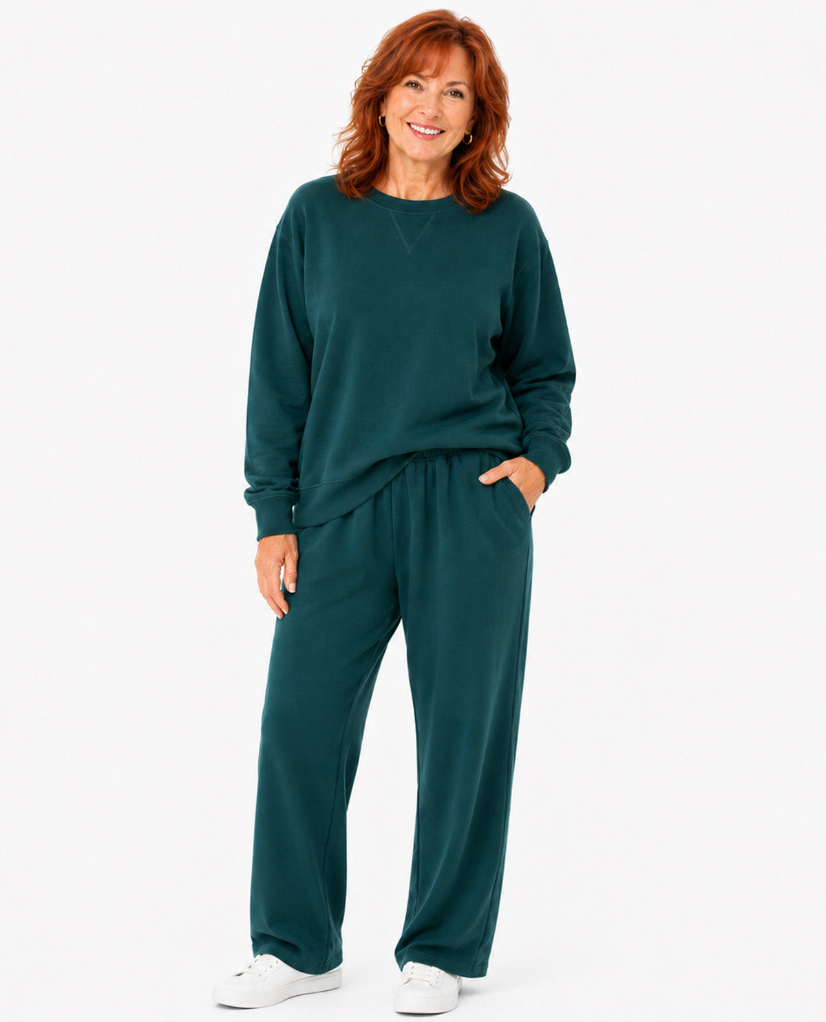 A woman modeling a matching dark teal green lounge set consisting of a long-sleeve sweatshirt and wide-leg sweatpants, paired with white sneakers, against a plain white studio background.