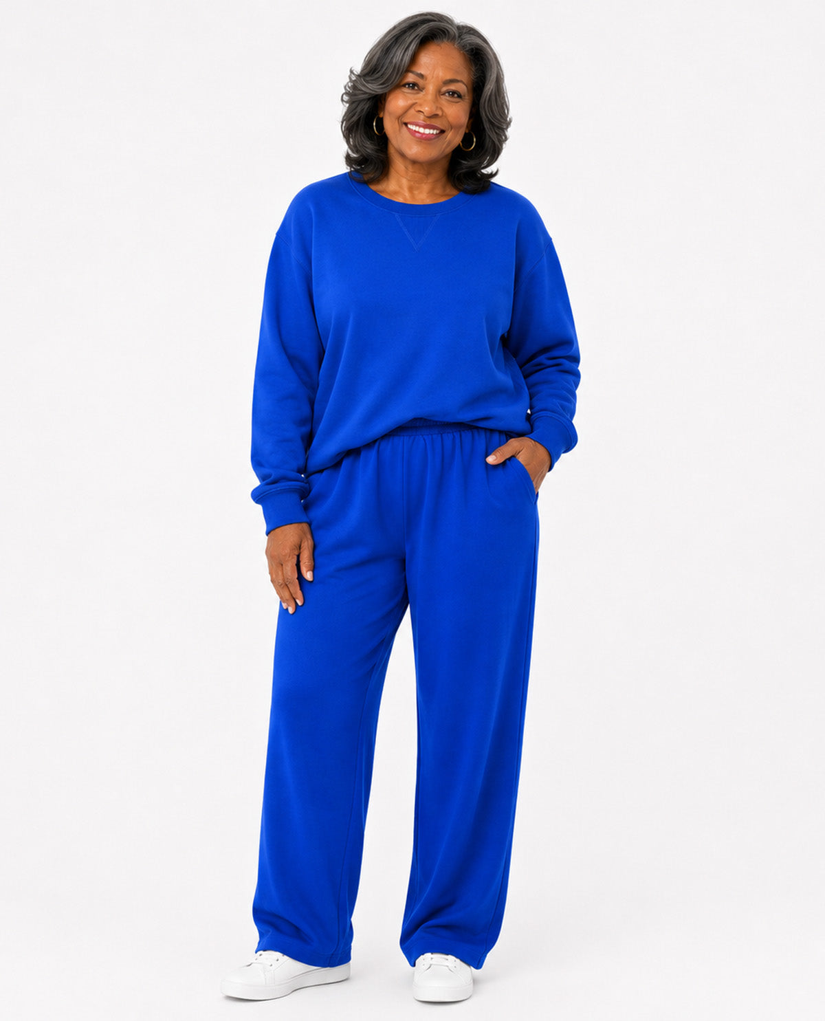 A smiling woman wearing a matching royal blue sweatshirt and wide-leg sweatpants set, paired with white sneakers, against a plain white studio background.