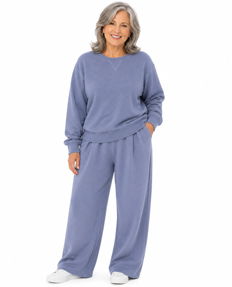 An older woman with gray hair stands against a white background wearing a matching dusty blue lounge set consisting of a long-sleeve sweatshirt and wide-leg pants.