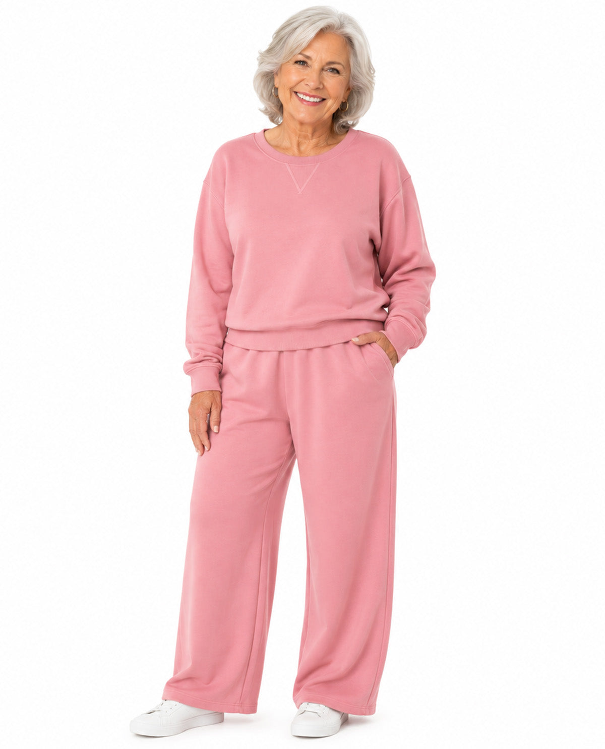 An older woman with short gray hair is modeling a matching pink lounge set consisting of a long-sleeve crewneck sweatshirt and wide-leg sweatpants, paired with white sneakers against a solid white background.