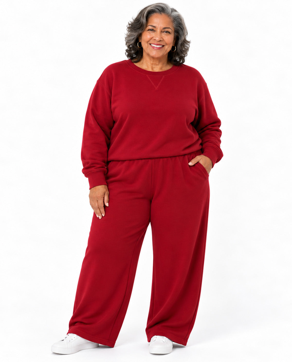 A smiling woman with gray hair wearing a matching red sweatshirt and wide-leg sweatpants set, paired with white sneakers, standing against a plain white background.